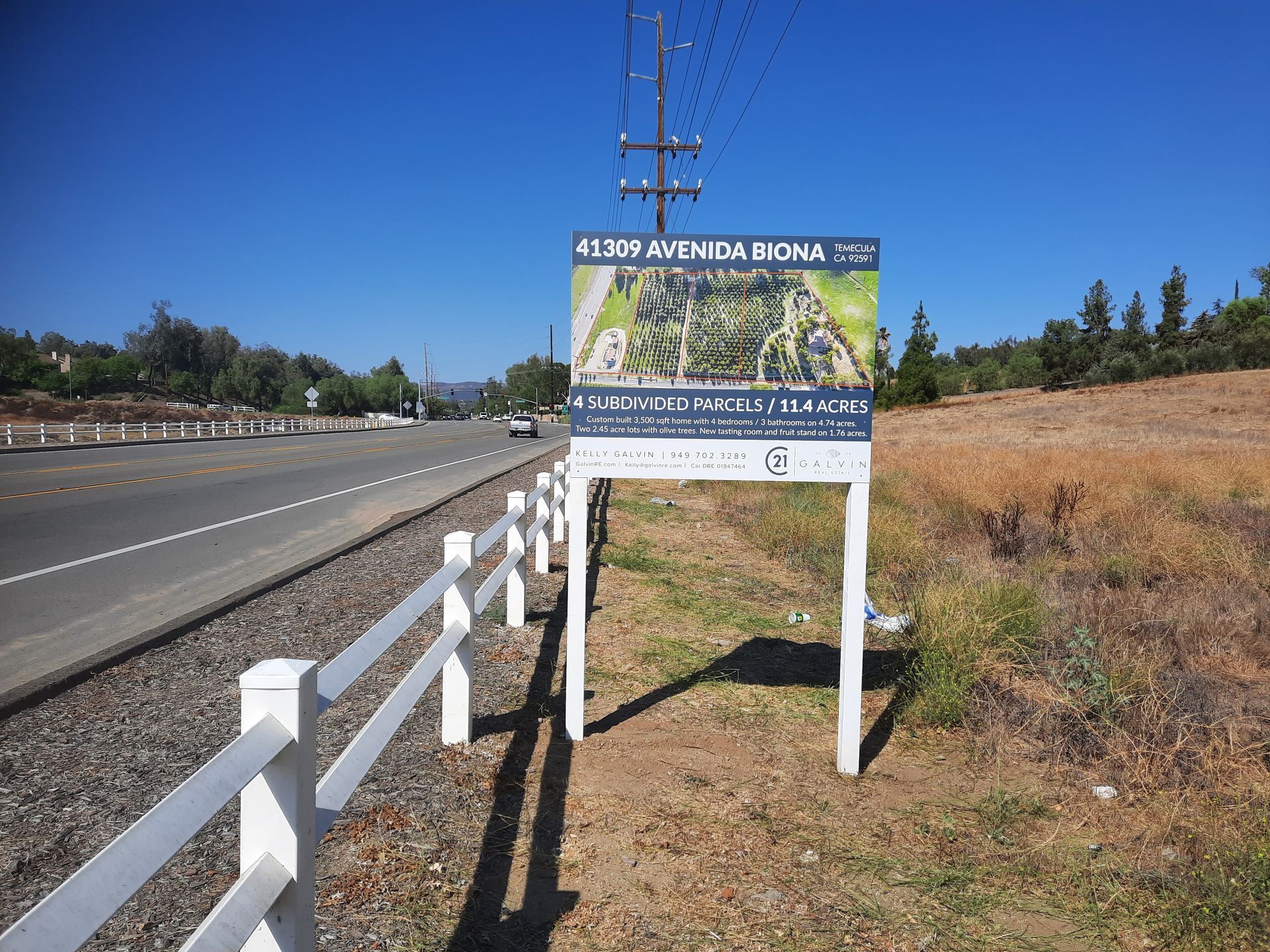 Roadside sign beside a highway, with white fence and grassy field under clear sky