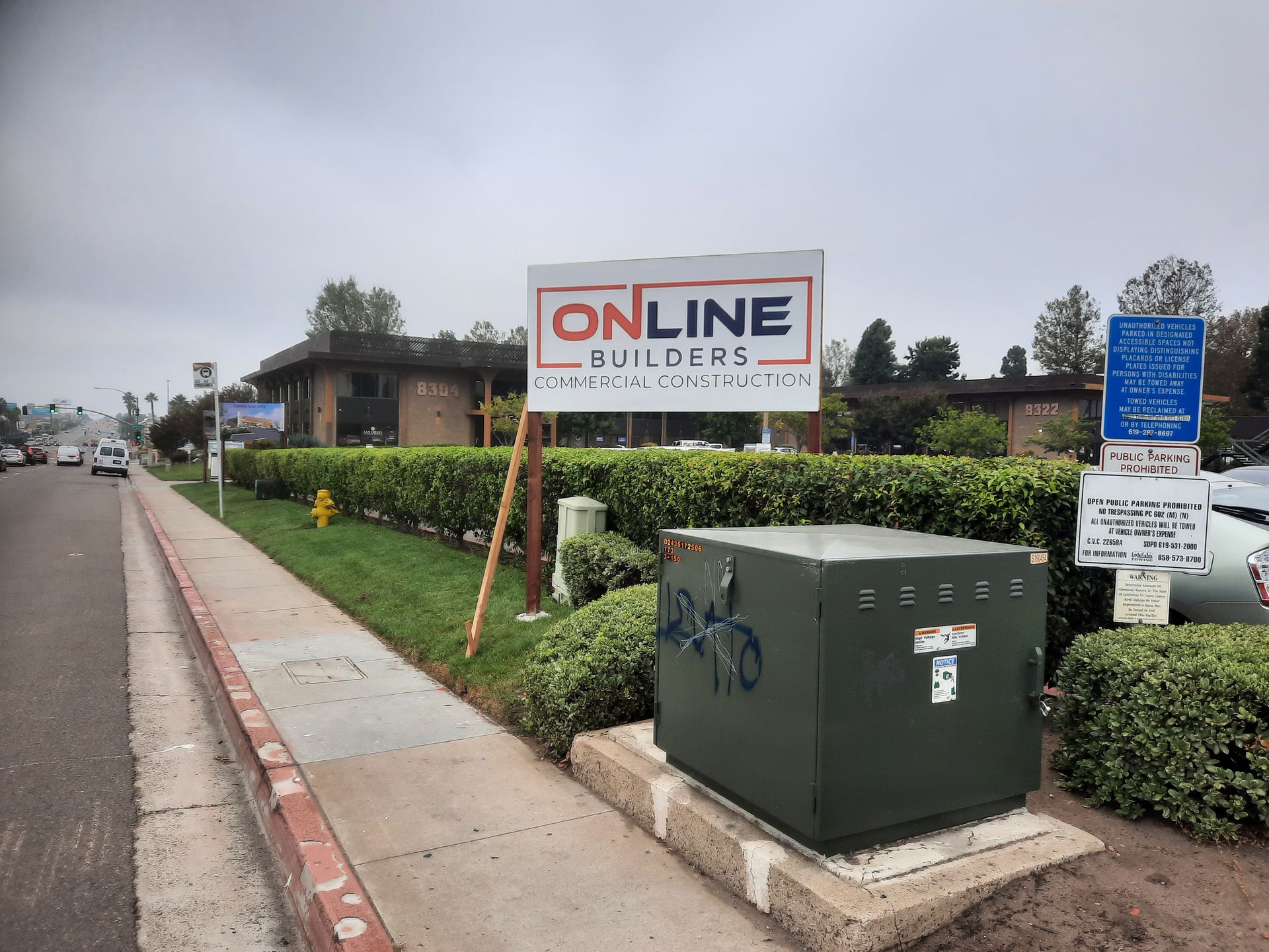Roadside sign for Online Builders beside a sidewalk and hedge, with utility boxes in front.
