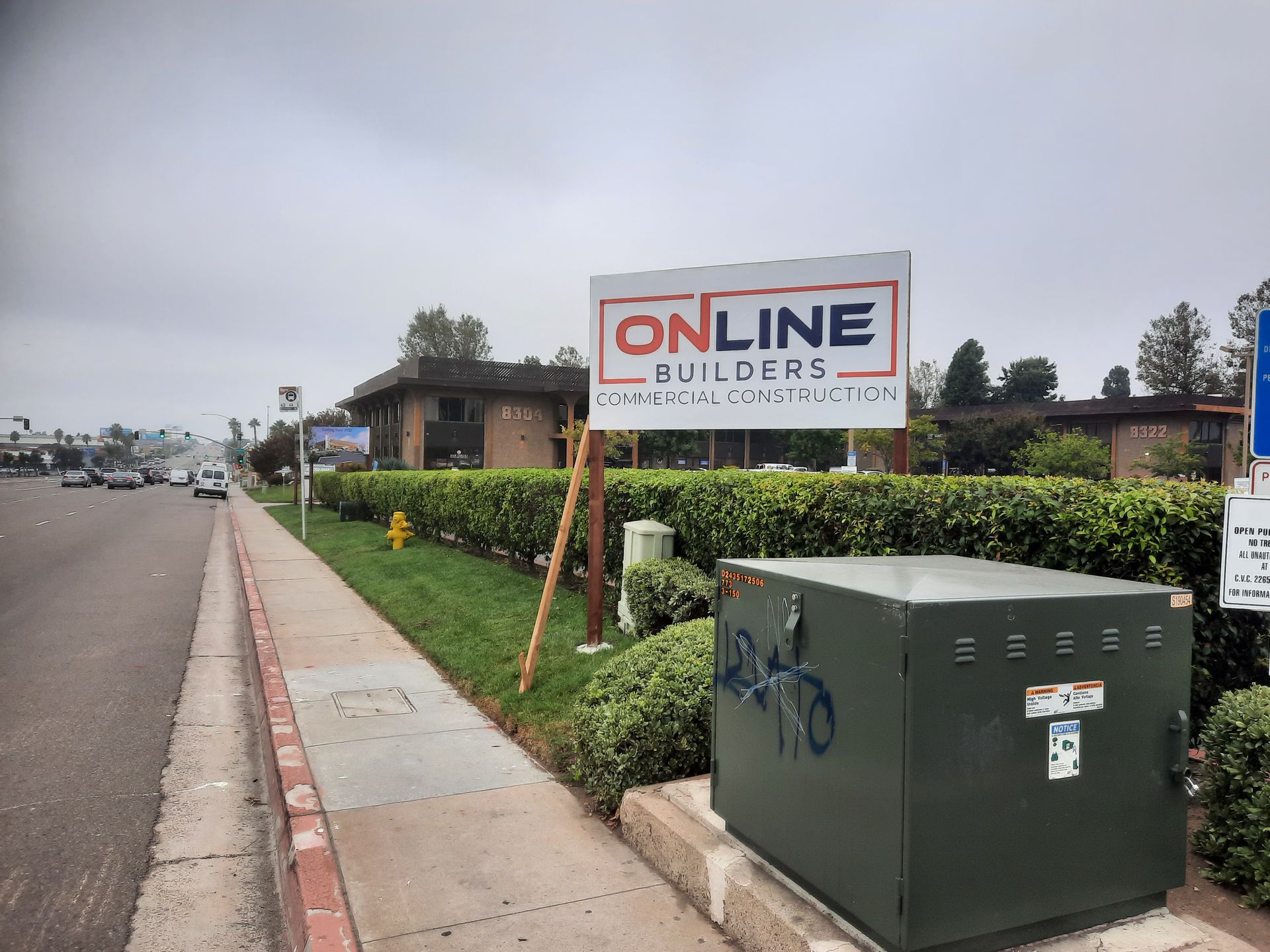 Street view with an “Online Builders” sign, hedge, utility box, and sidewalk beside a roadway.
