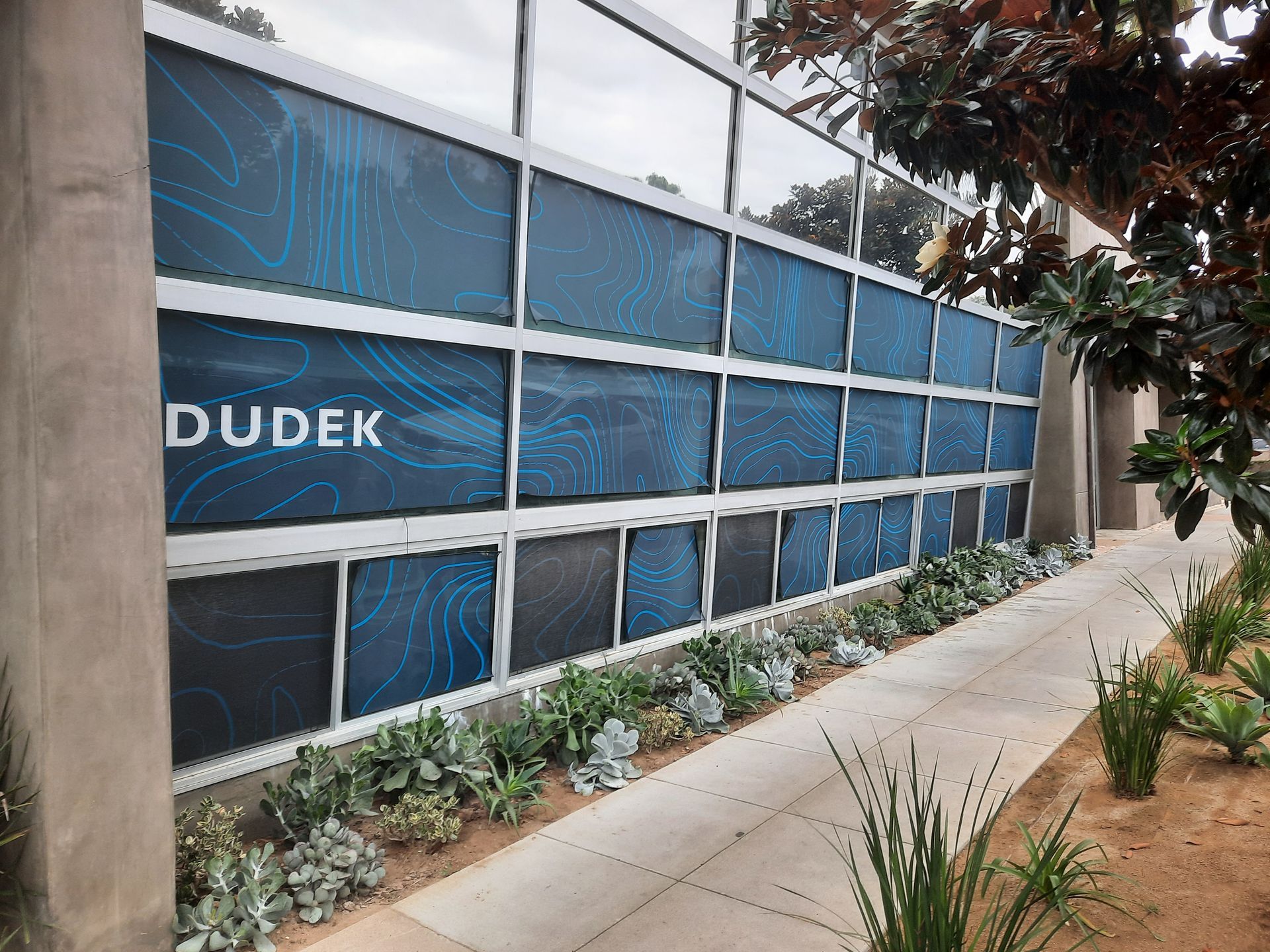 Blue glass windowed building facade with the word “DUDEK” and a landscaped sidewalk in front