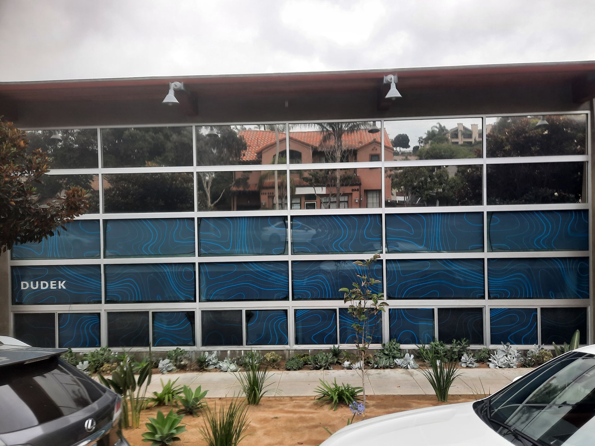 Exterior of a modern building with blue glass windows, parked cars, and a small landscaped entrance