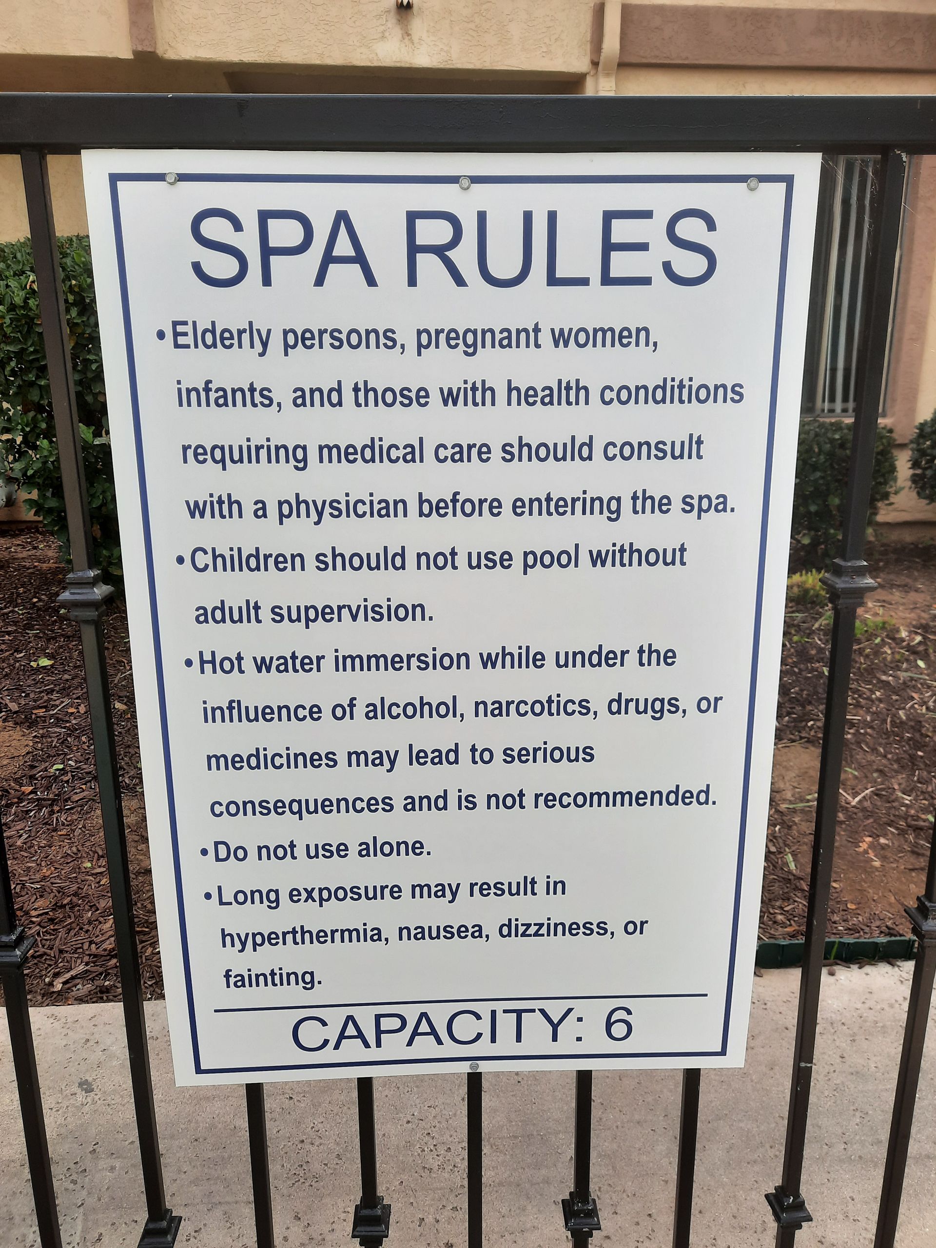 Spa rules sign on a black metal gate, white background with blue border, listing spa safety guidelines and capacity 6