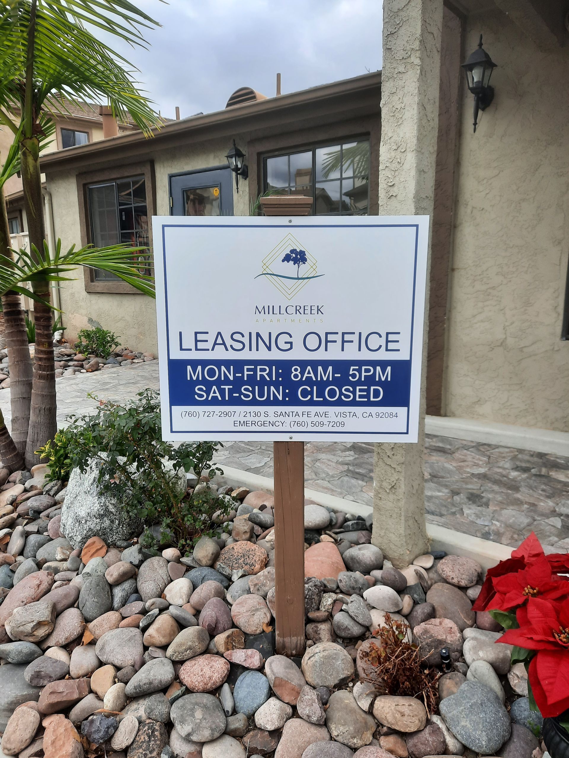 Leasing office sign outside a building, with hours listed and rocks and flowers in the foreground