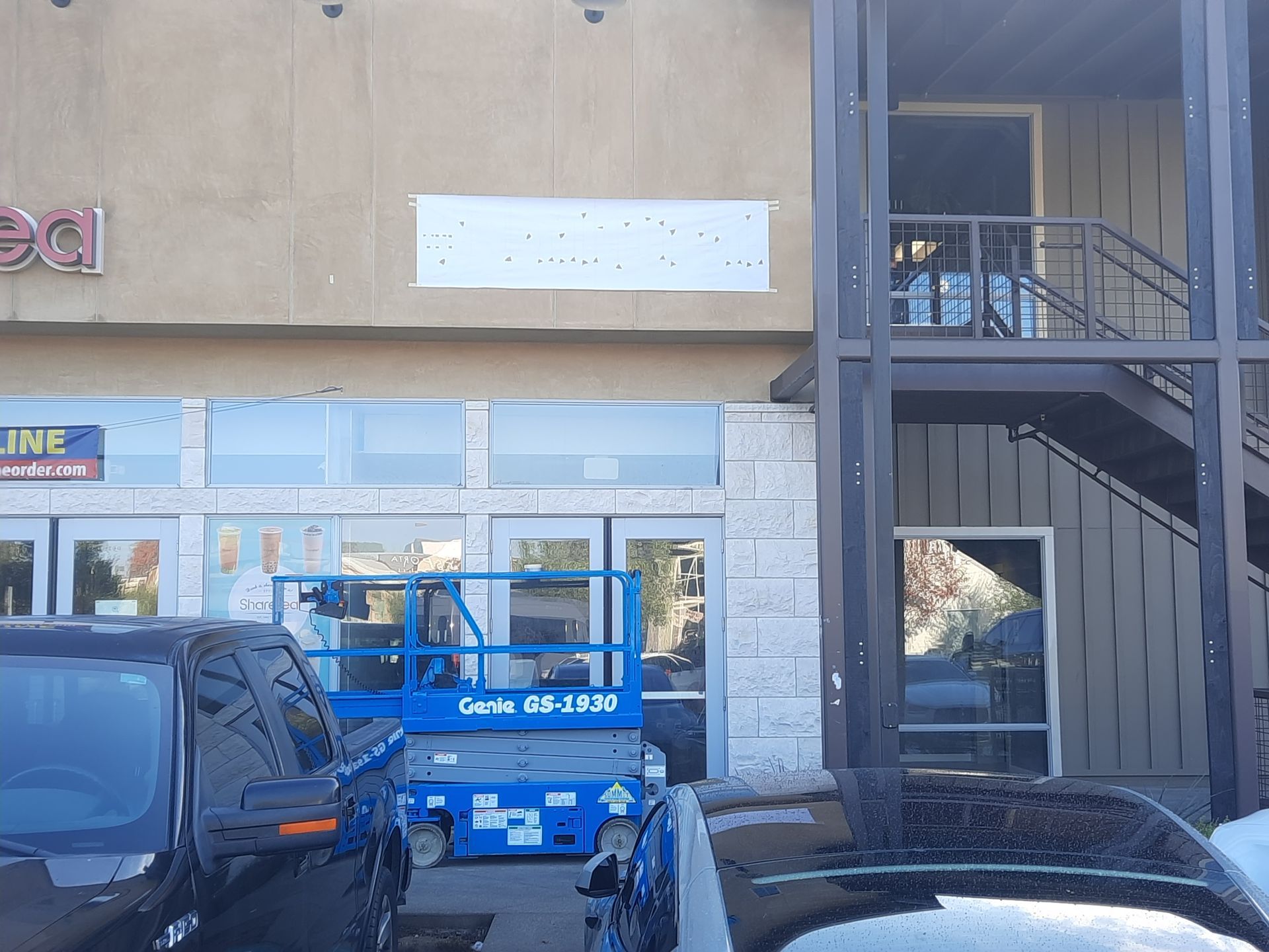 Blue cherry picker parked in front of a store entrance beside parked cars and a stairway