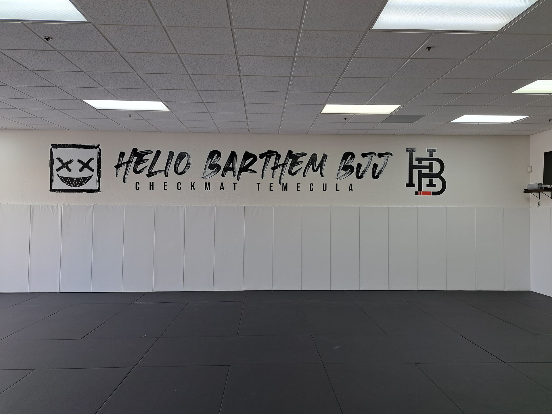 Empty martial arts studio with black mat floor and a wall logo reading “Hello Spartan BJJ”