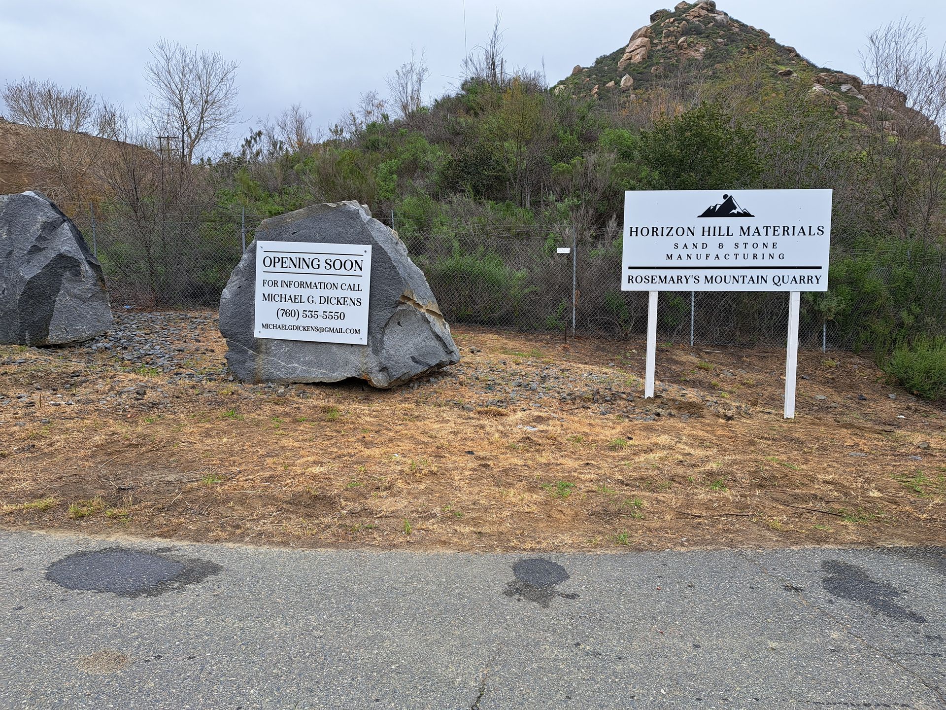 Trailhead signs beside a rocky path with dry brush and a hill in the background