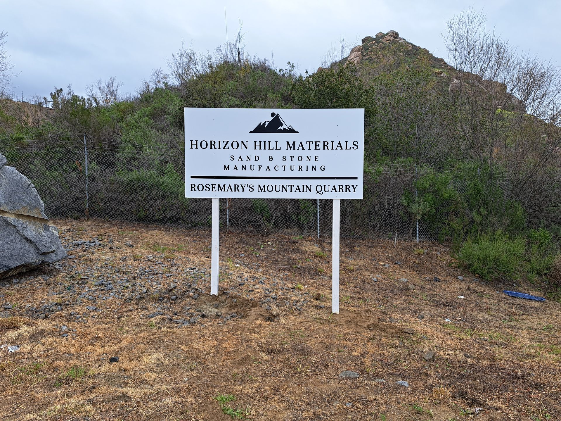 White sign for Horizon Herb Watering on posts beside a dirt trail in rocky, brushy terrain.
