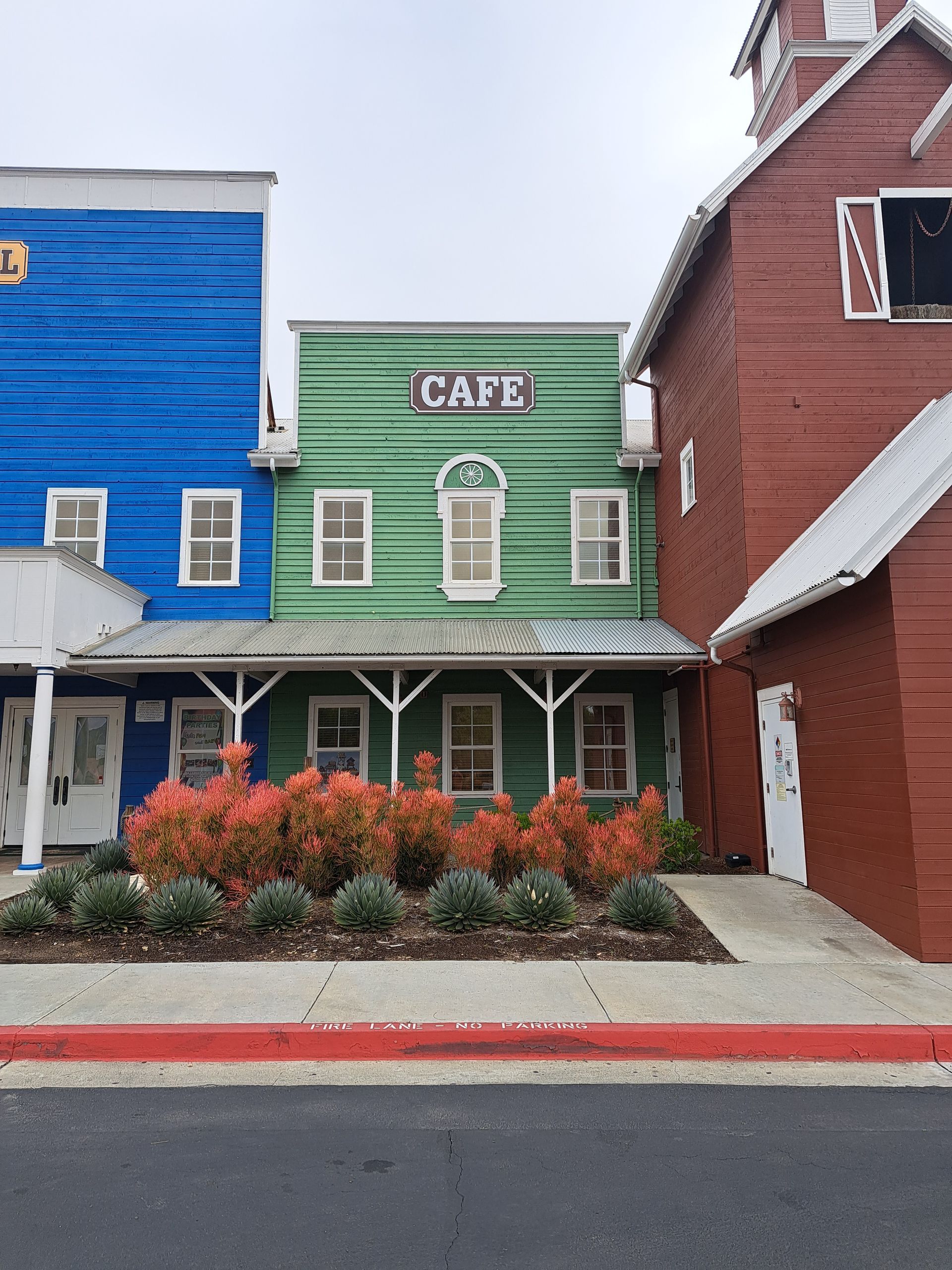 Small green cafe storefront between blue and red buildings, with a porch and flower beds.