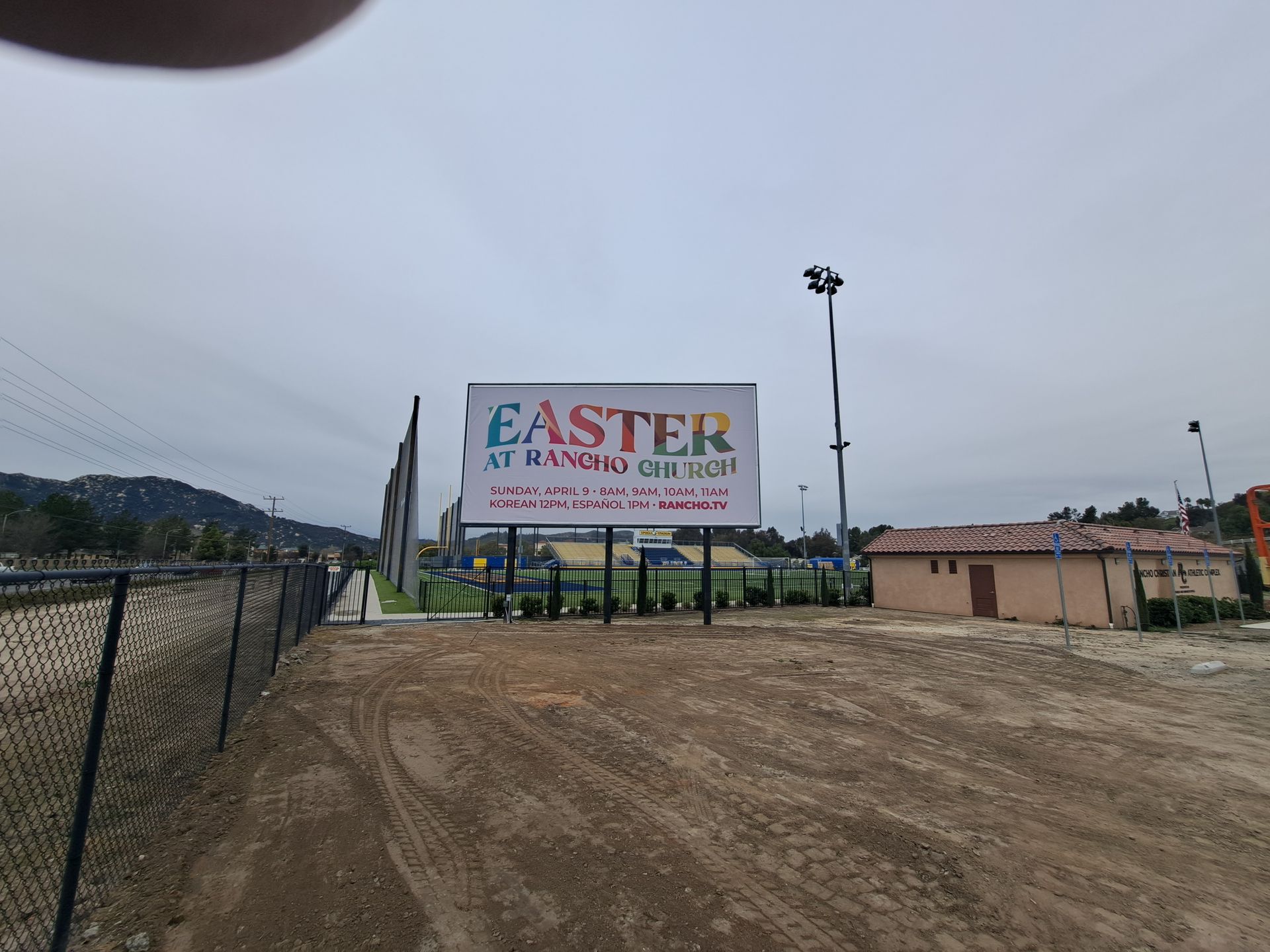 School signboard at a dirt lot: “Easter Kindergarten” under a cloudy sky.