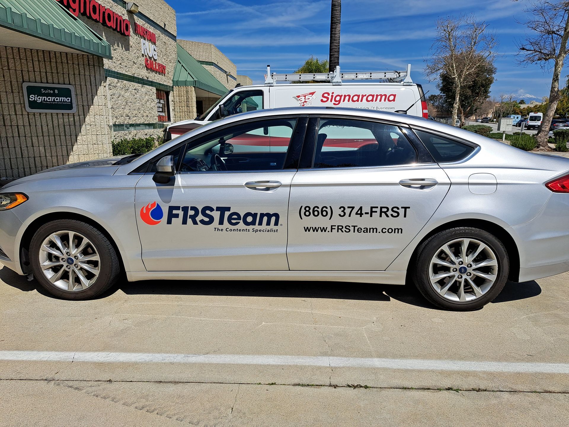 White FIRST Team sedan with company branding parked outside a storefront on a sunny street.