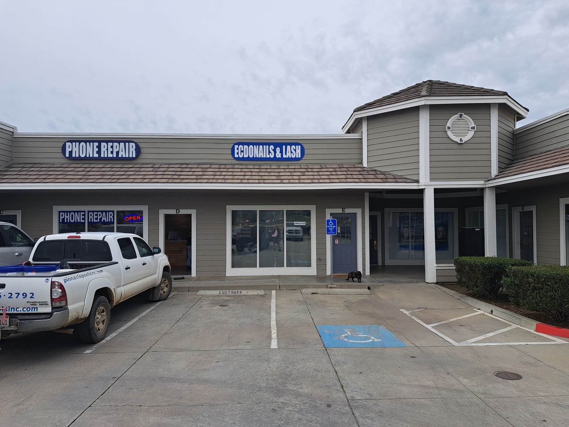 Strip mall storefronts with a parked white pickup and accessible parking spaces under an overcast sky