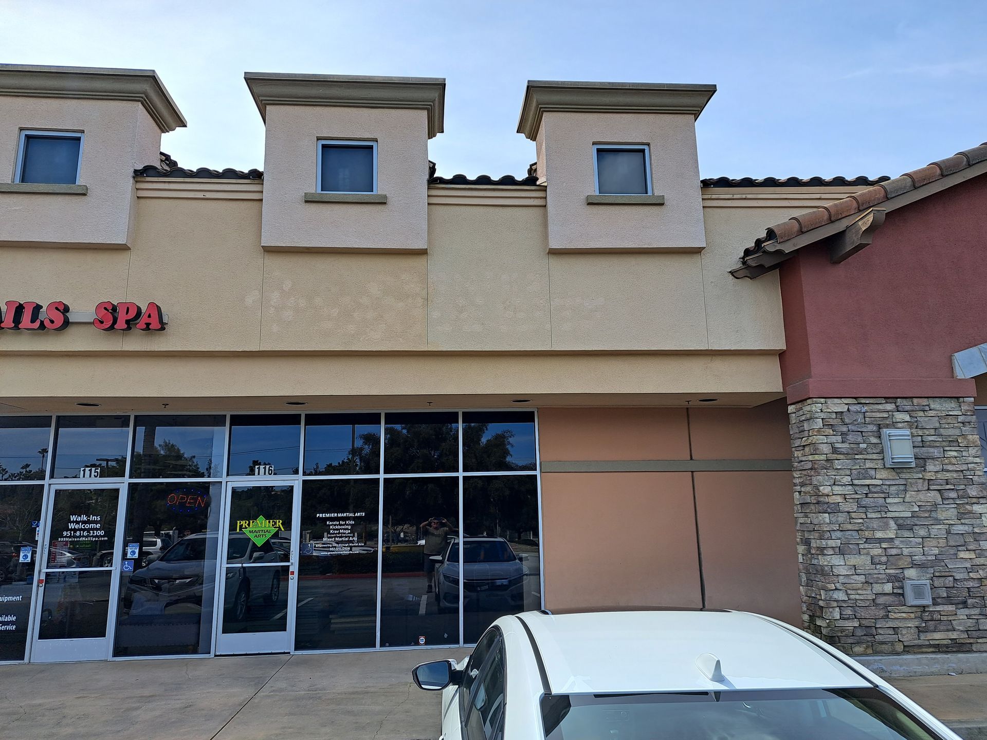 Strip mall storefront with tan stucco facade, glass doors, stone wall, and a white car parked in front.