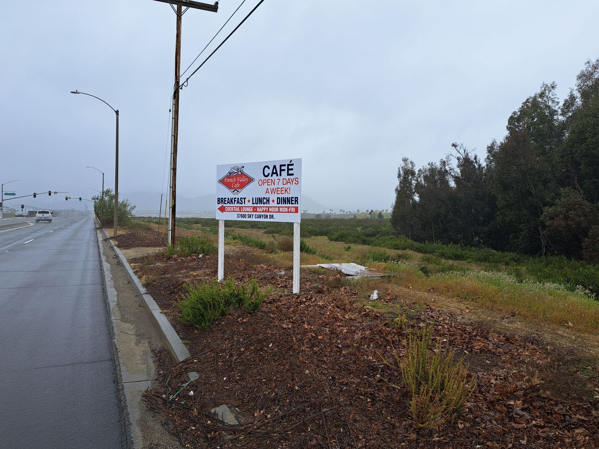 Roadside café sign beside a wet rural road under a cloudy sky