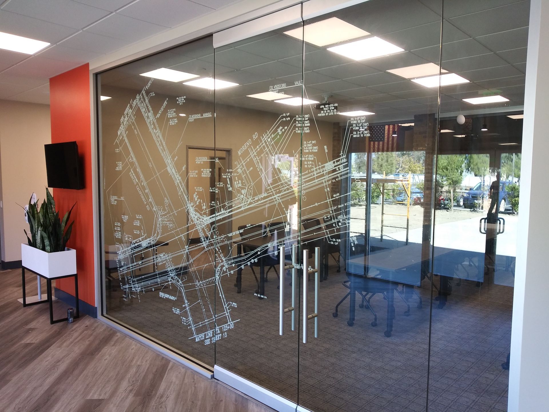 Glass office entrance with etched pattern, wooden floor, and potted plant near a wall-mounted TV.