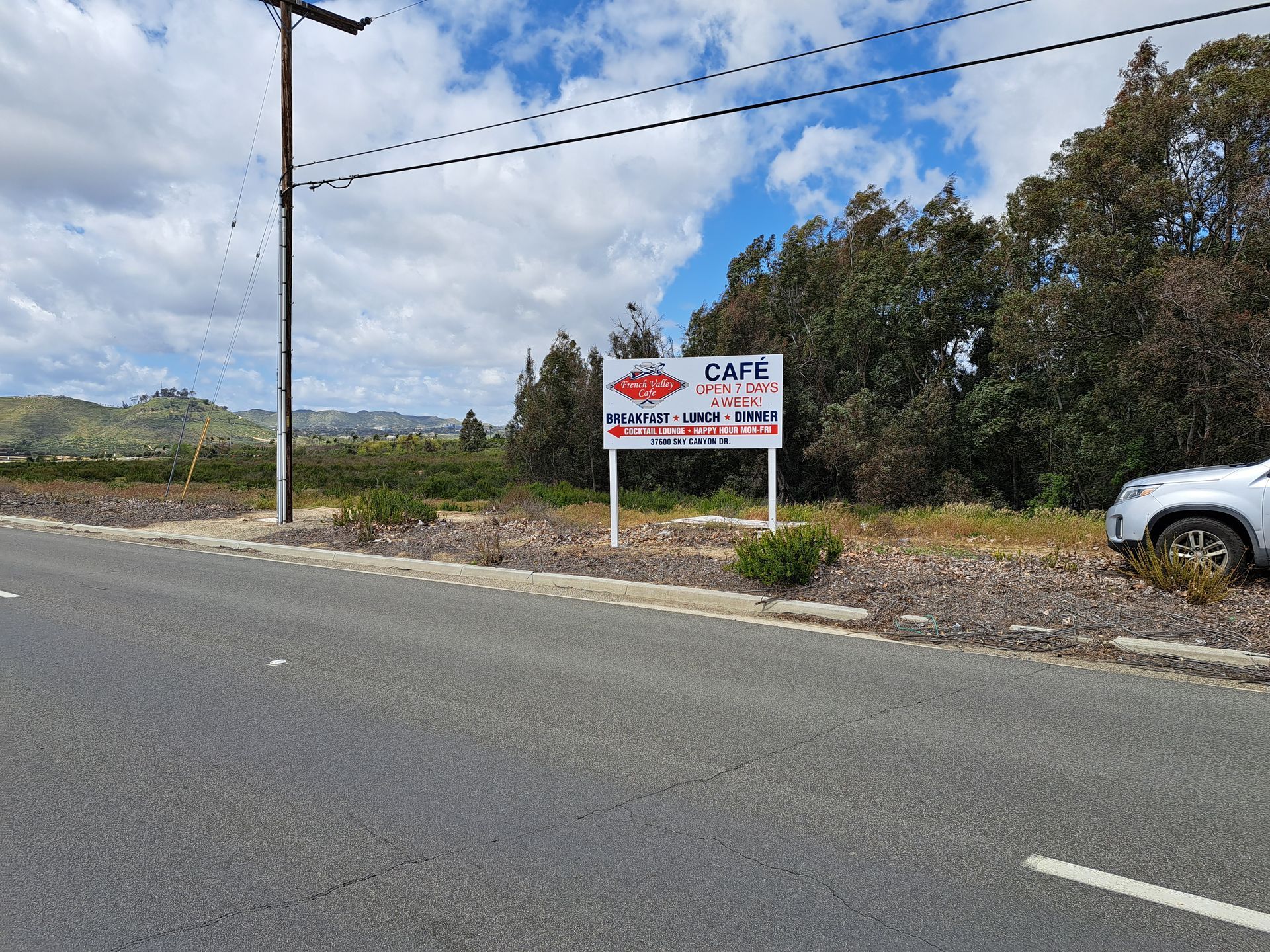 Roadside sign for a café near a rural highway, with trees, hills, and a parked white vehicle nearby.