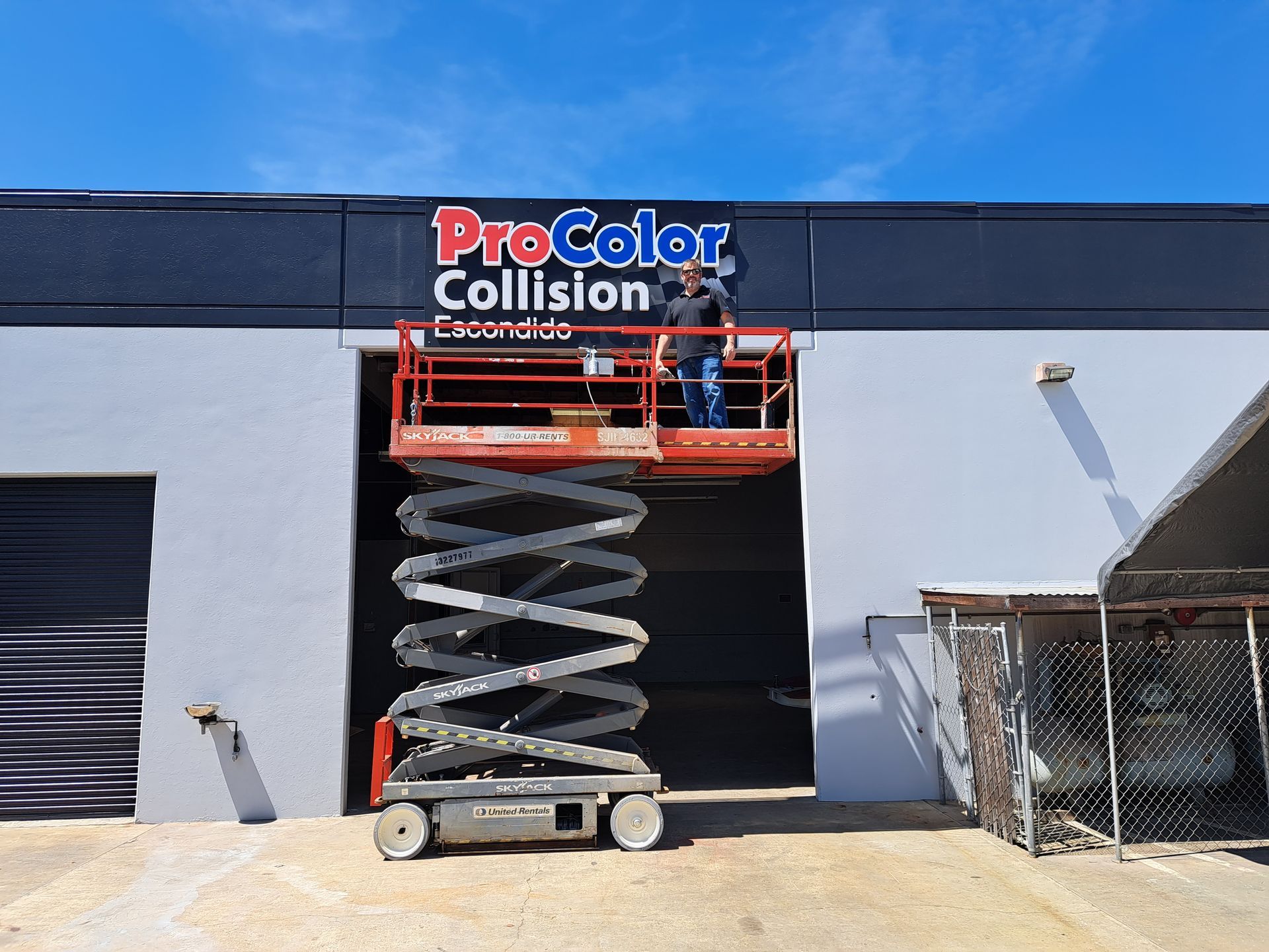 Scissor lift raised outside Pro Color Collision auto body shop under a clear blue sky