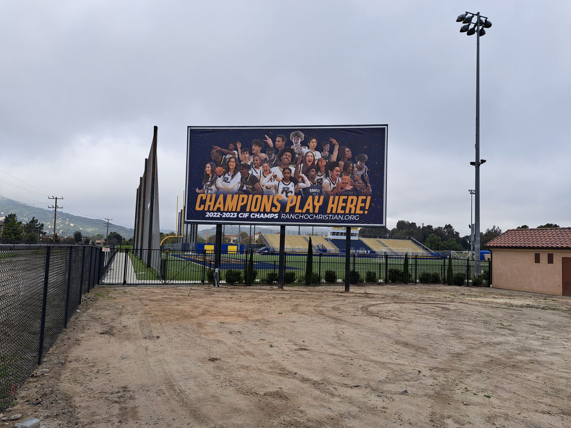 Baseball field entrance with a large “Champions Play Here” sign and stadium lights under a cloudy sky
