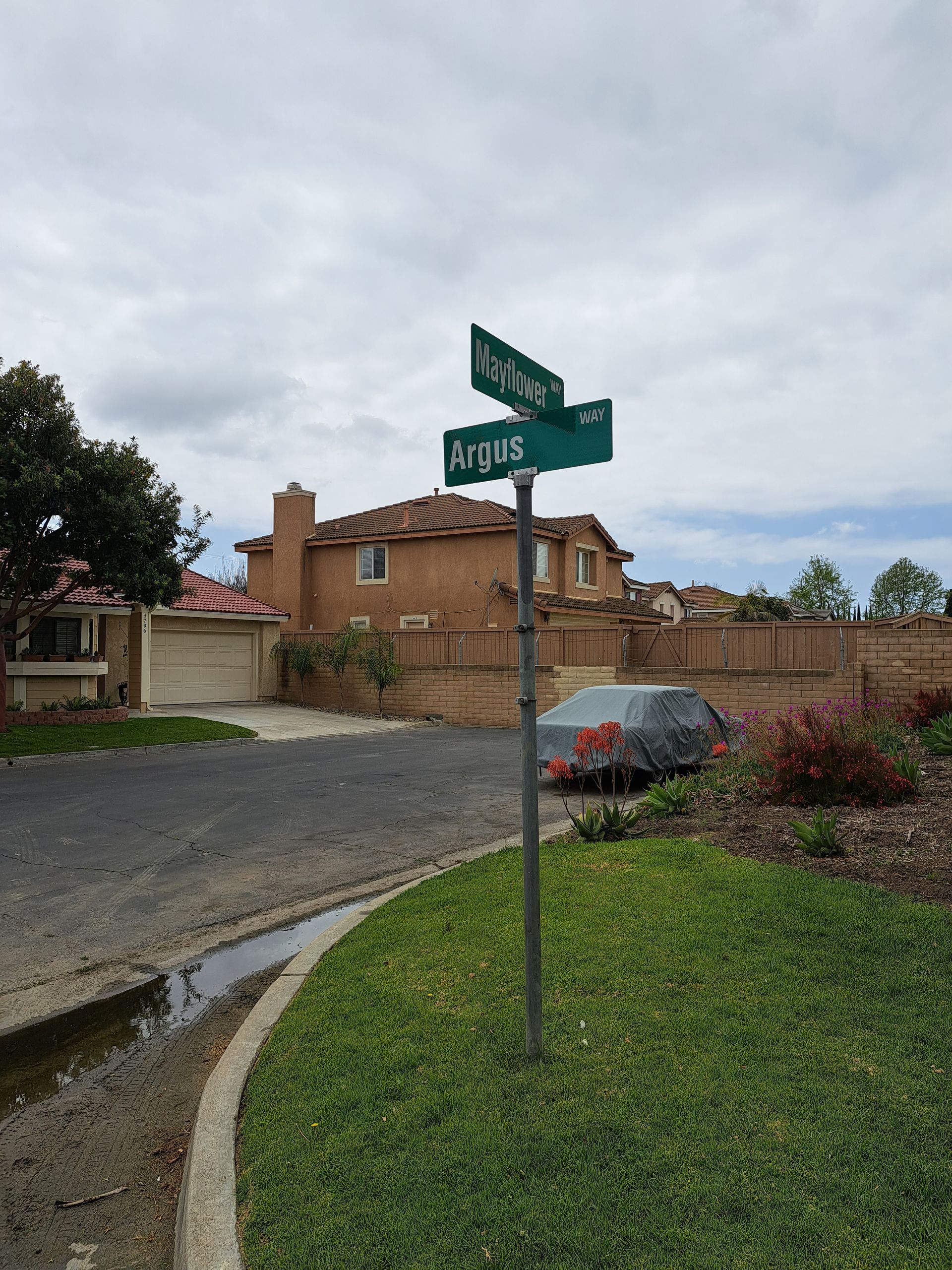 Street signs at a rural intersection beside a brick house, parked car, and overcast sky.