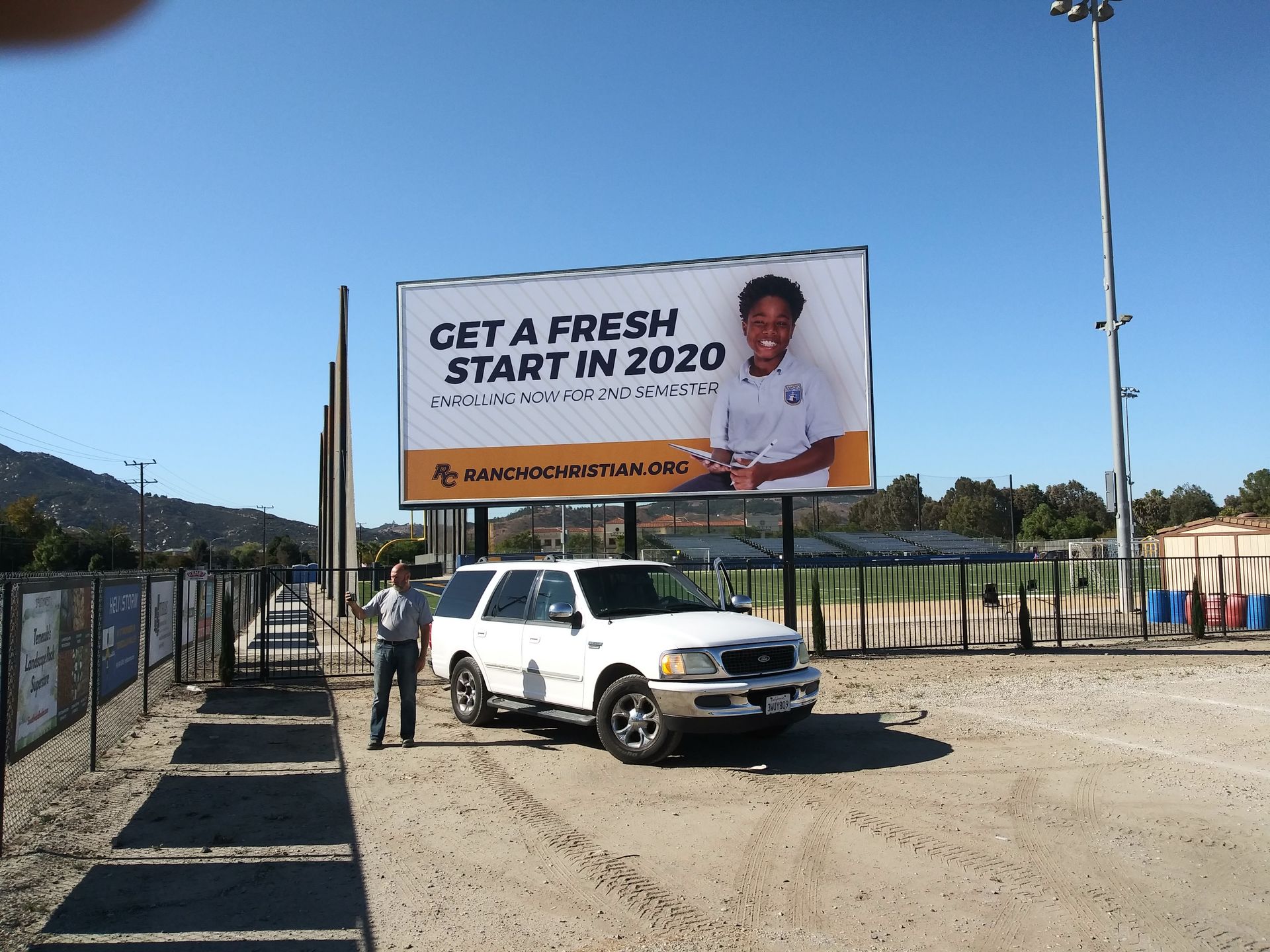 White pickup truck under a billboard on a dirt road at a sunny outdoor event entrance