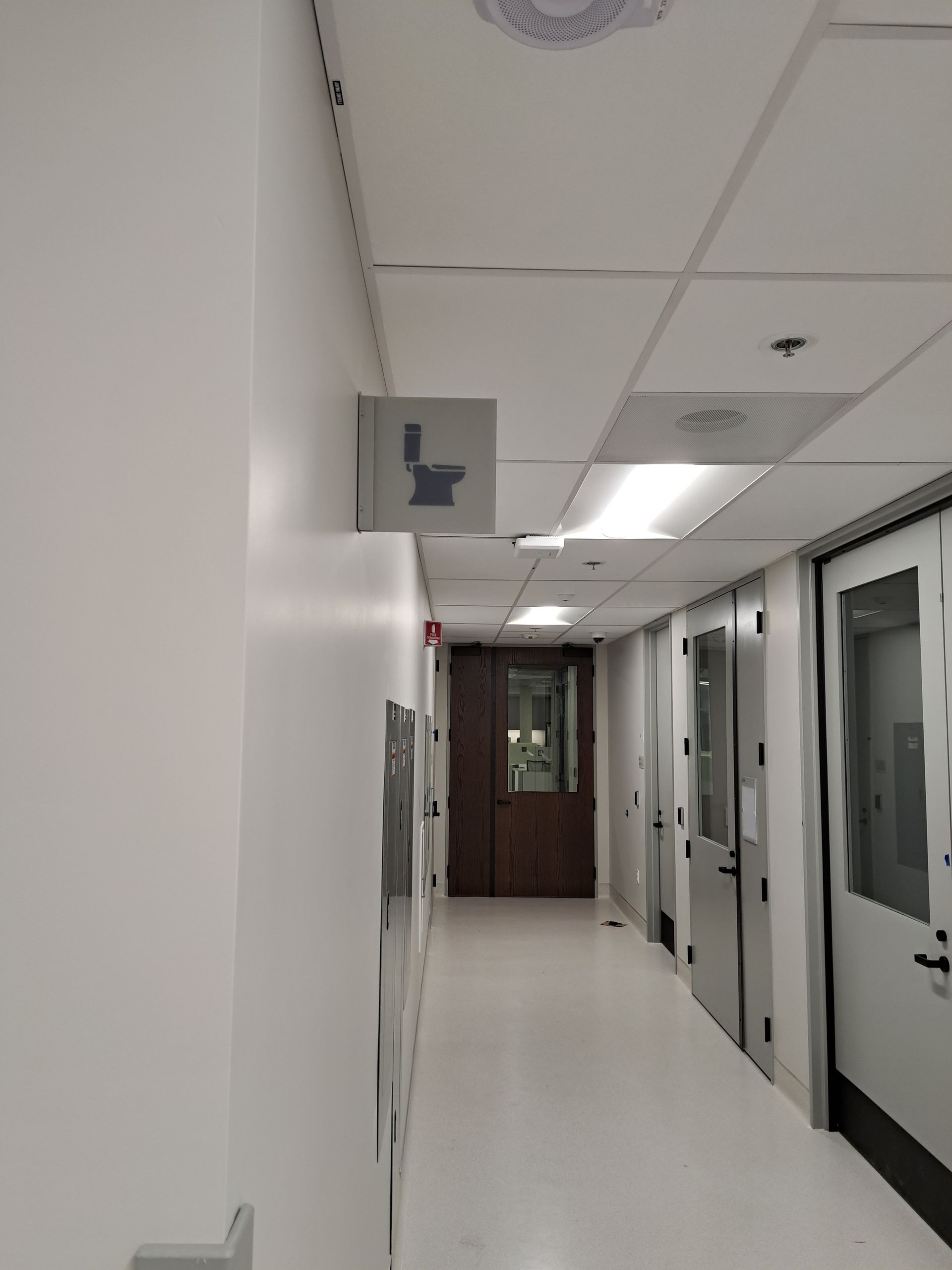 Empty hospital hallway with white walls, glass doors, and a sign hanging from the ceiling.