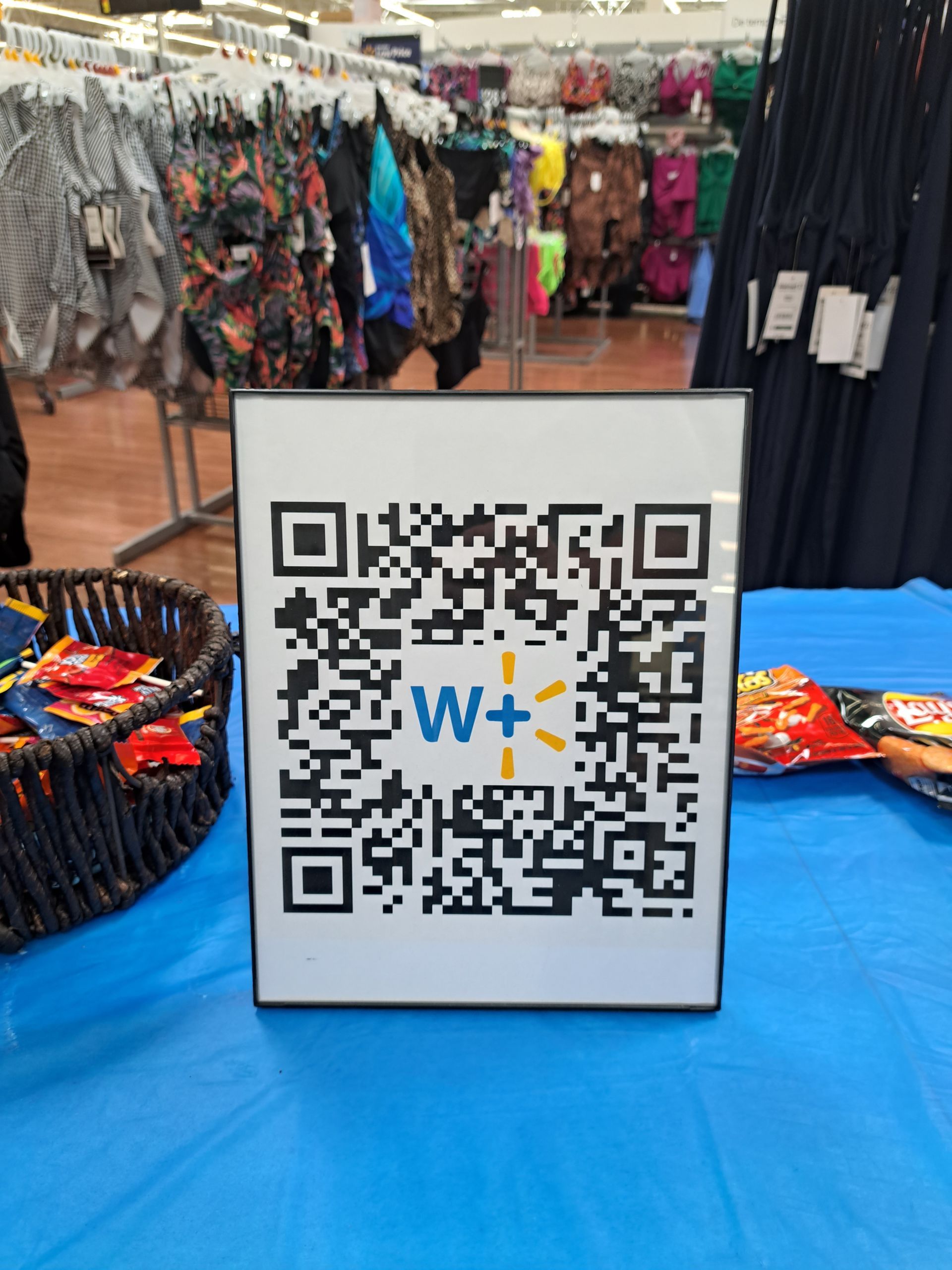 QR code sign on blue table at a clothing market stall with racks of garments in the background