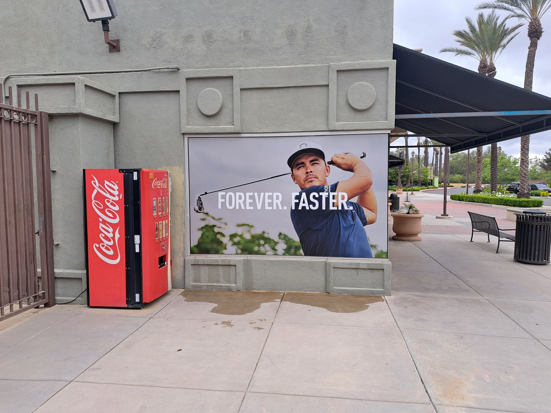 Coca-Cola vending machines beside a wall mural of a smiling person with “FOREVER FASTER” text outdoors