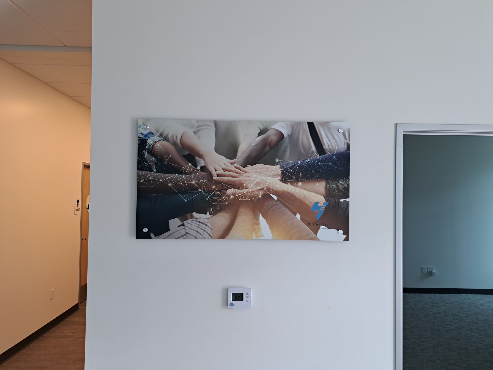 Hallway wall with a large photo of hands stacked together, beside an open doorway and thermostat.