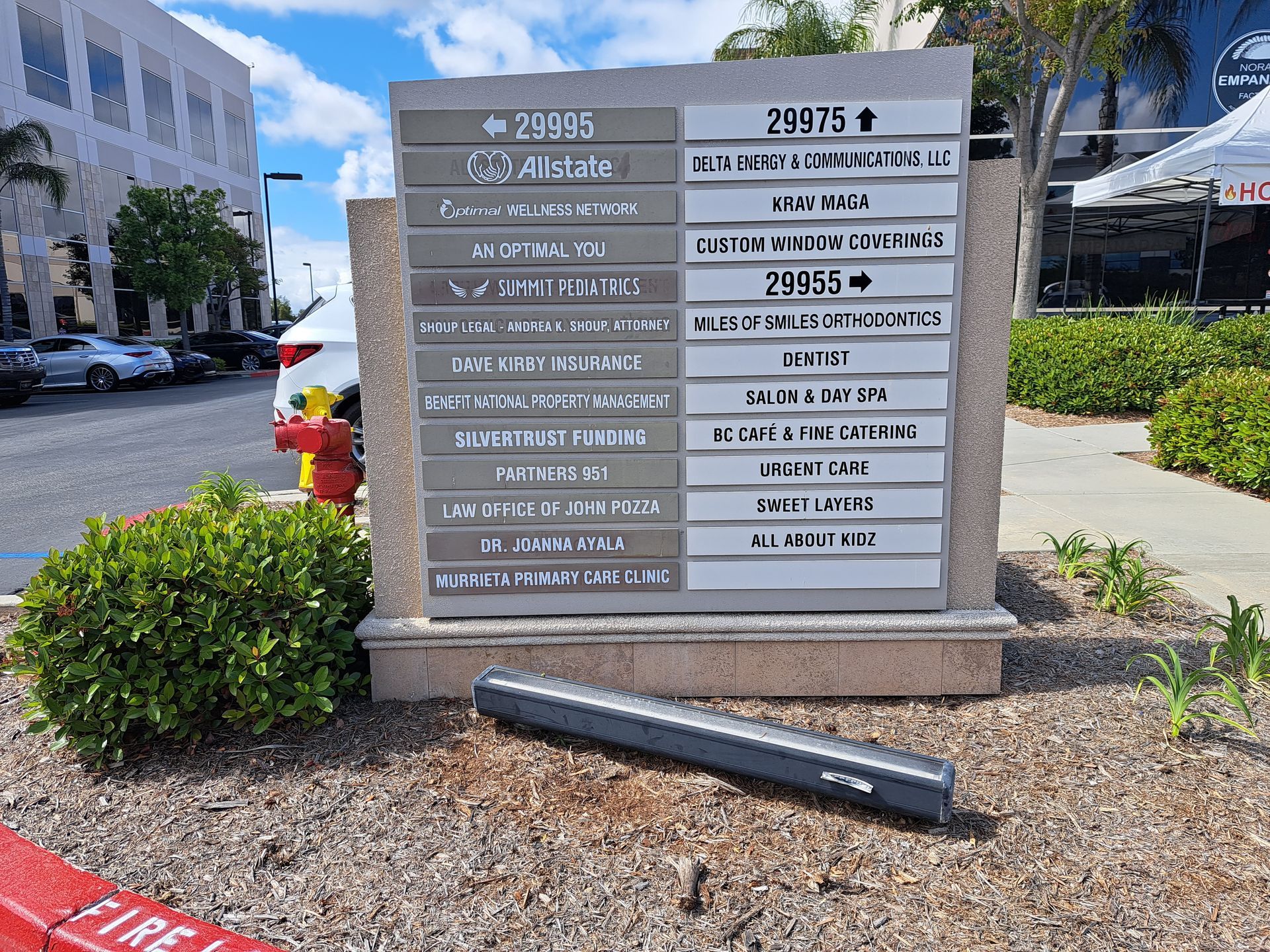 Directory sign outside a building on a sunny sidewalk, with landscaping and a fallen black pole in front.