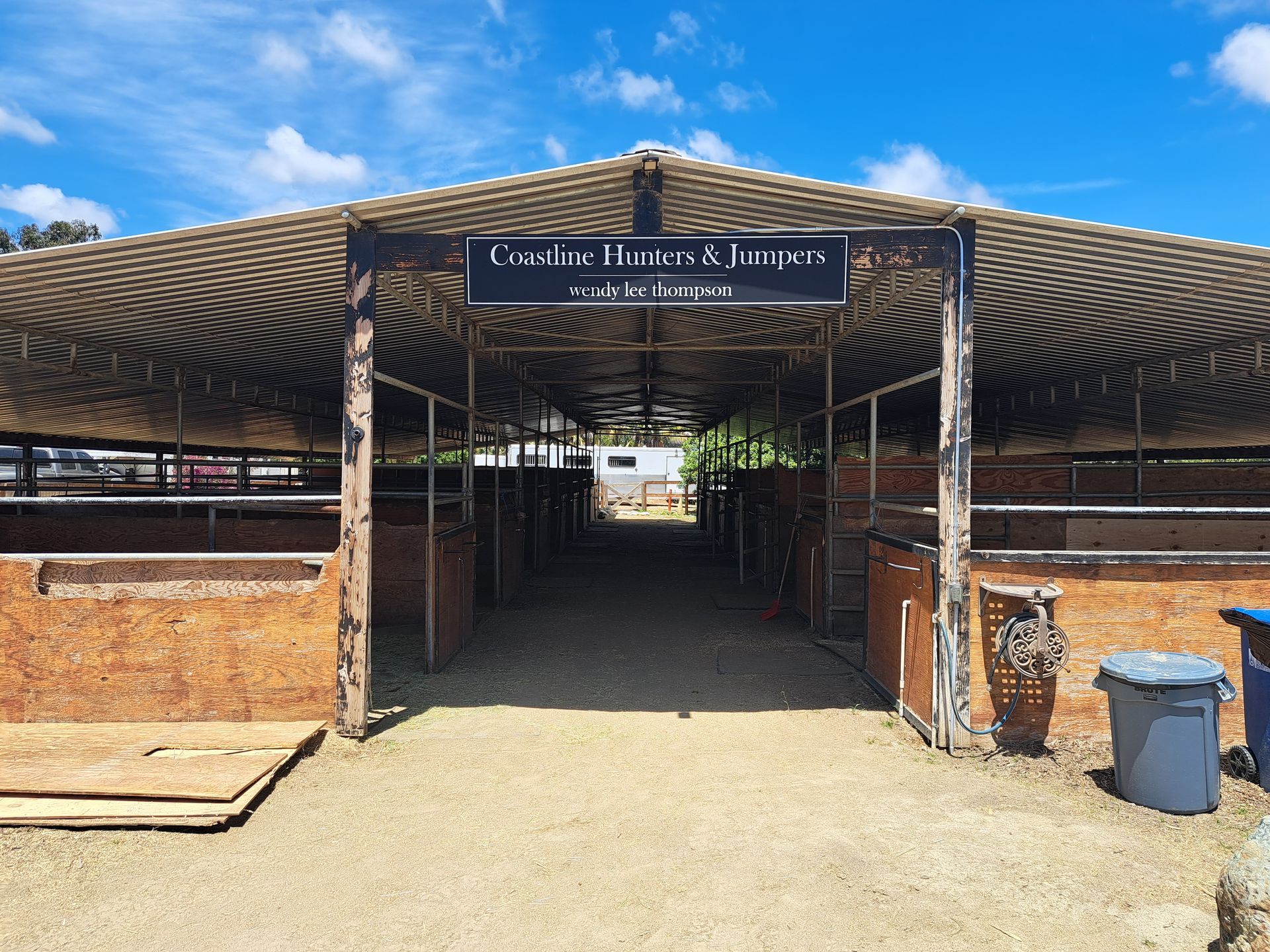 Open-air livestock pen with a sign, brick walls, and a sandy path under a striped canopy