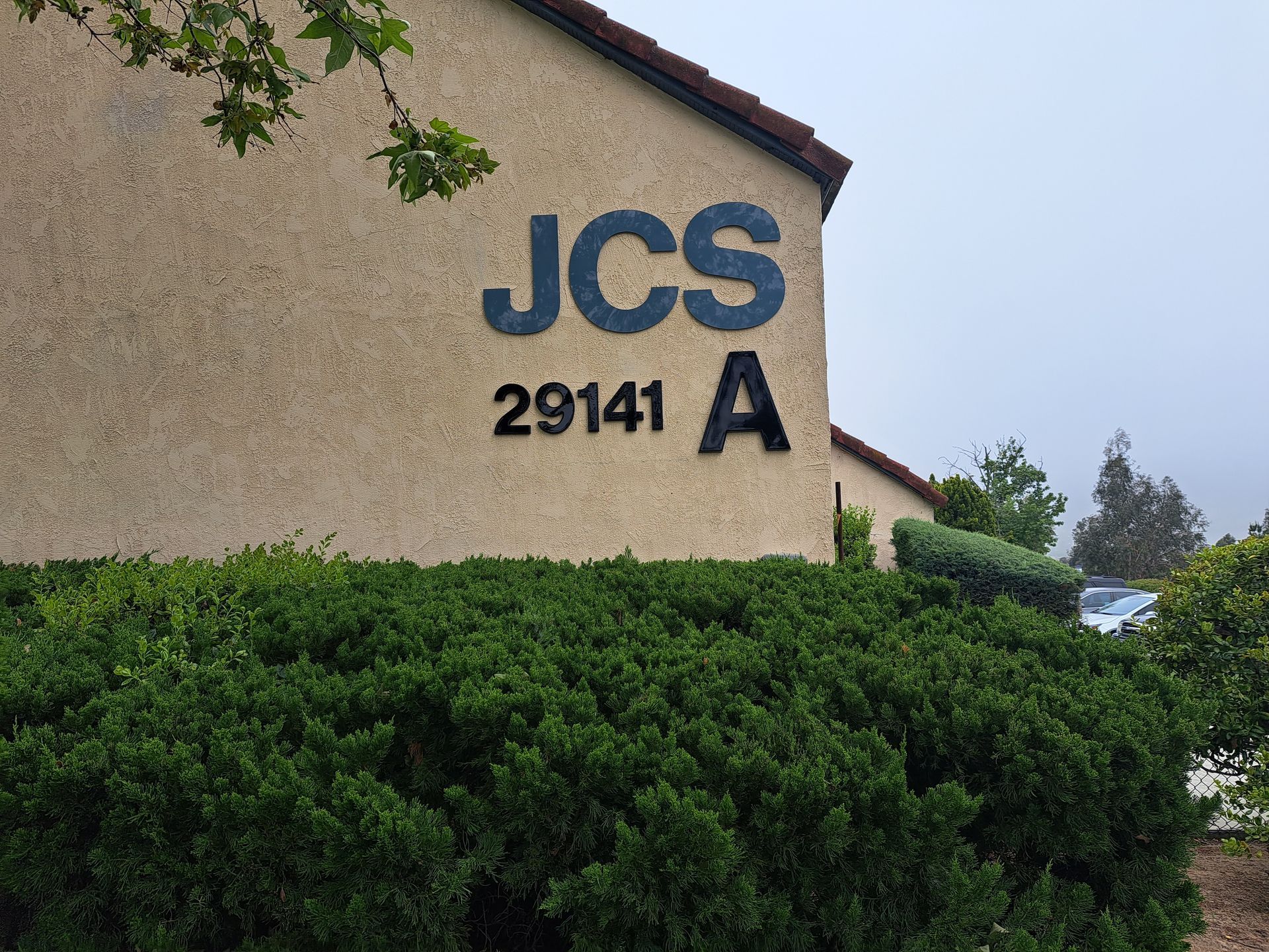 Building exterior with “JCS” and “2914 A” on a stucco wall above green hedges.