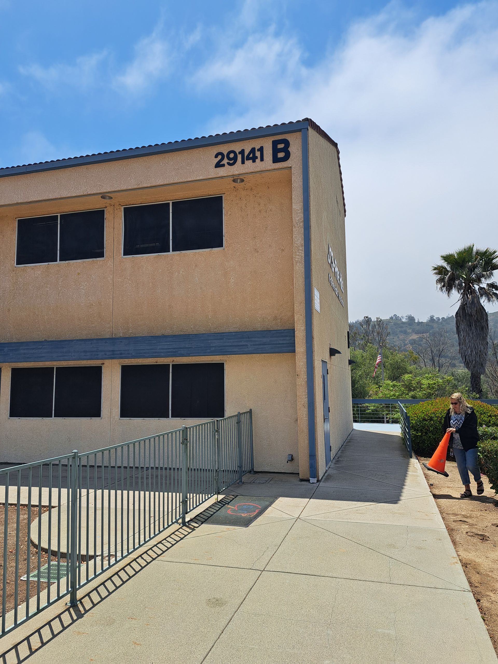 Two-story beige building with dark windows and a fenced walkway, with a person near the entrance.