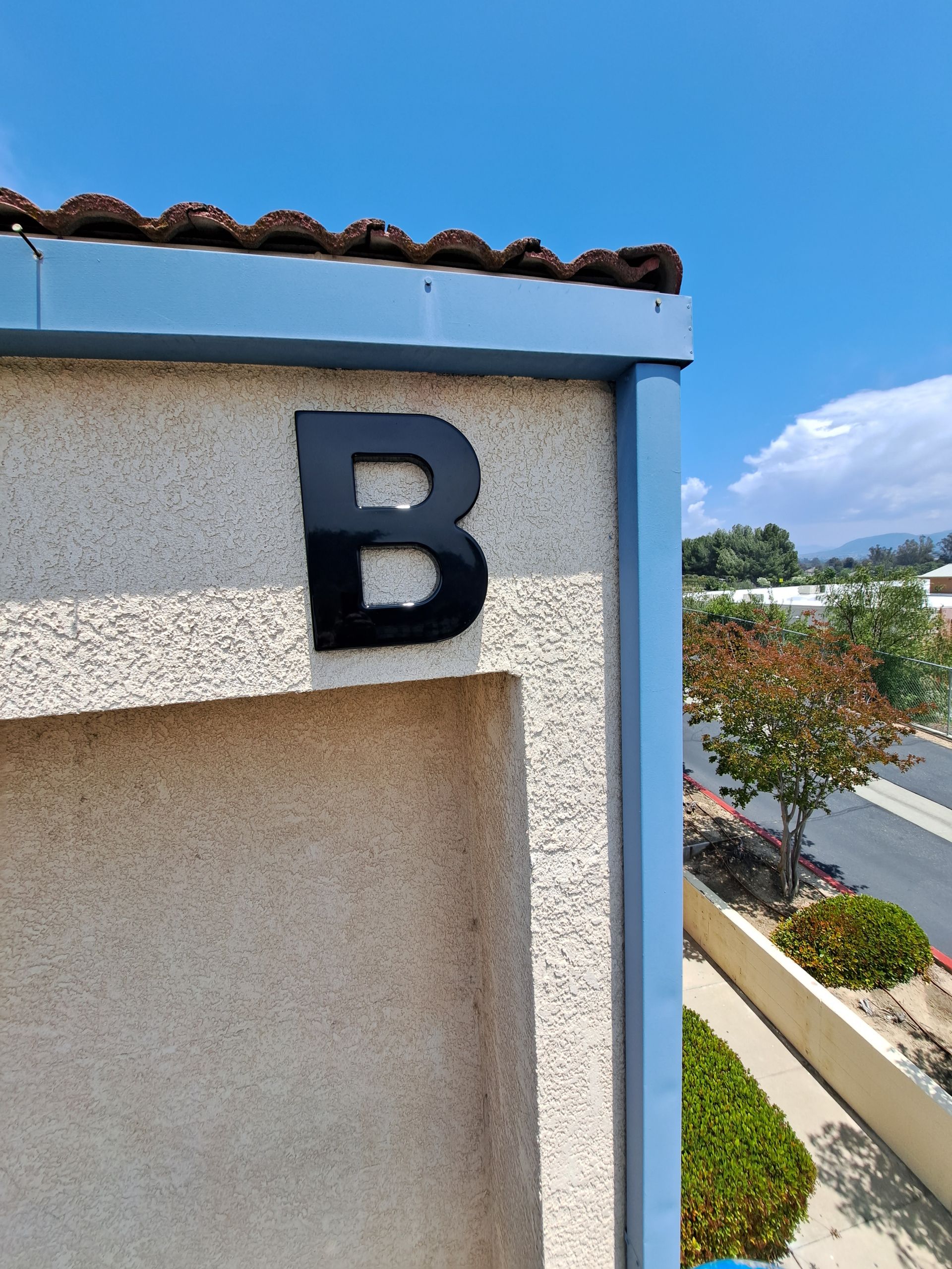 Black letter B on a stucco wall beside a blue-roofed building and road under a clear sky