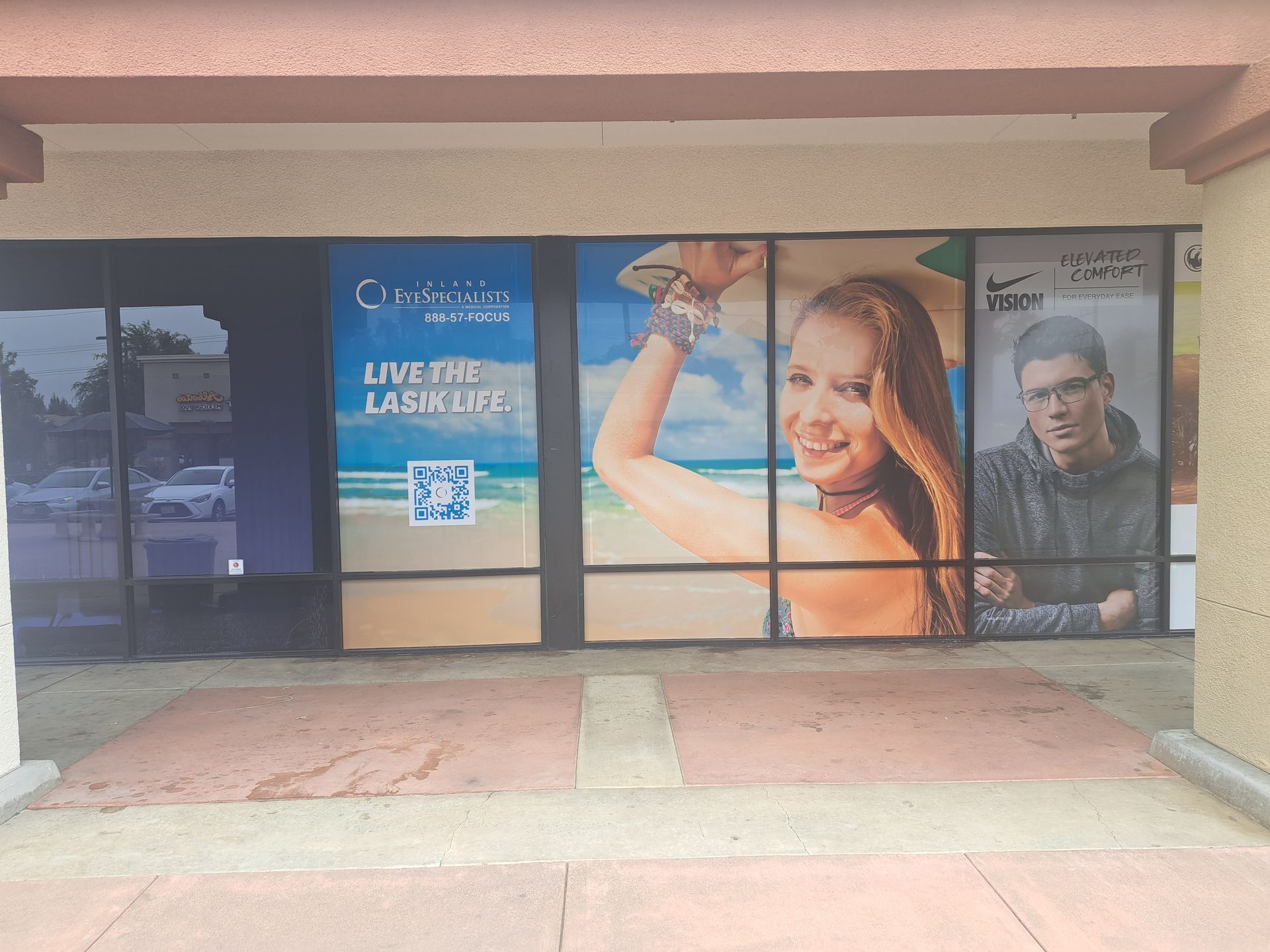 Storefront with large window ads, including a smiling woman and man, under a covered walkway.