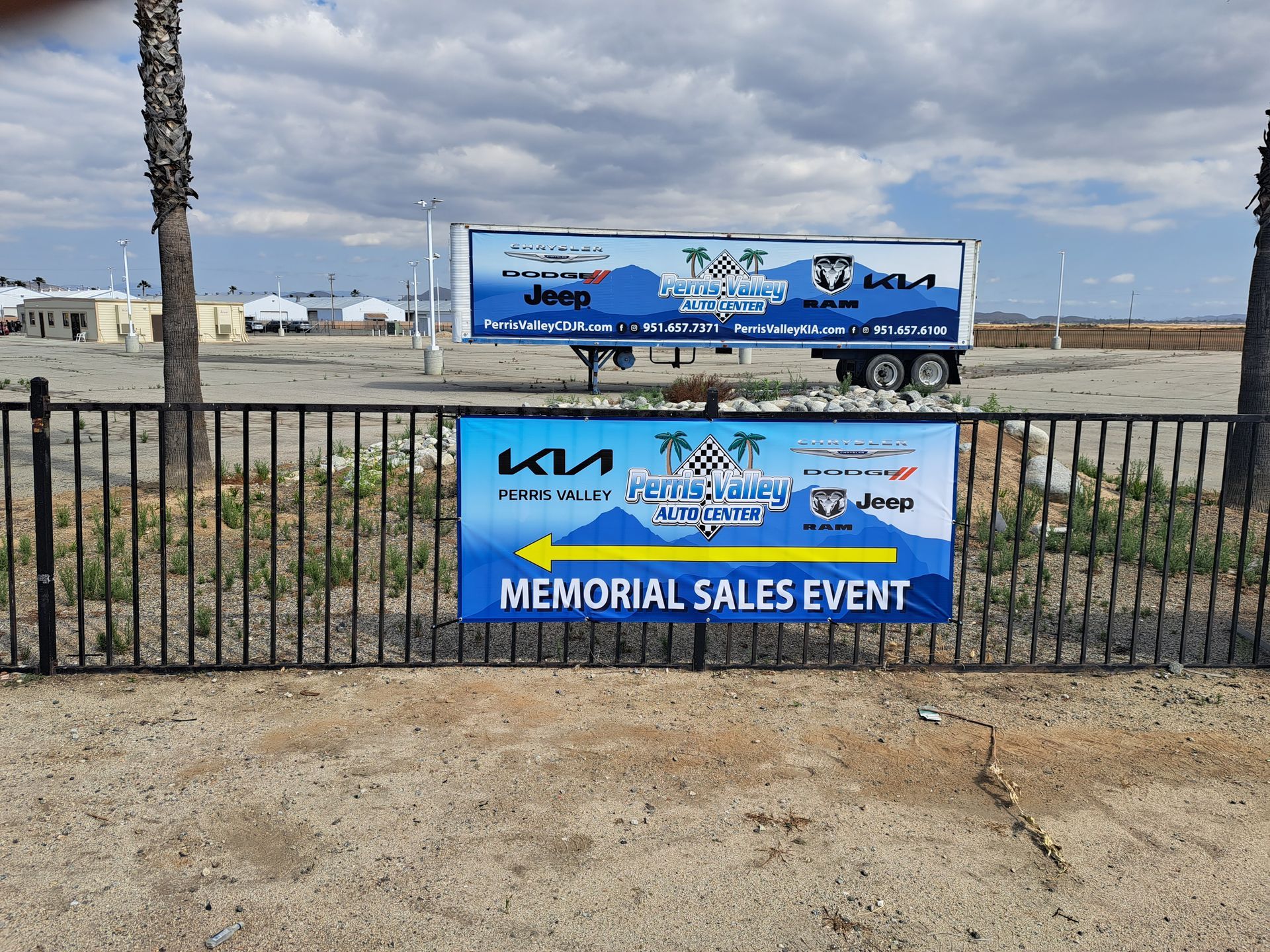 Memorial sales event sign on a fence with a blue trailer and cloudy sky behind it