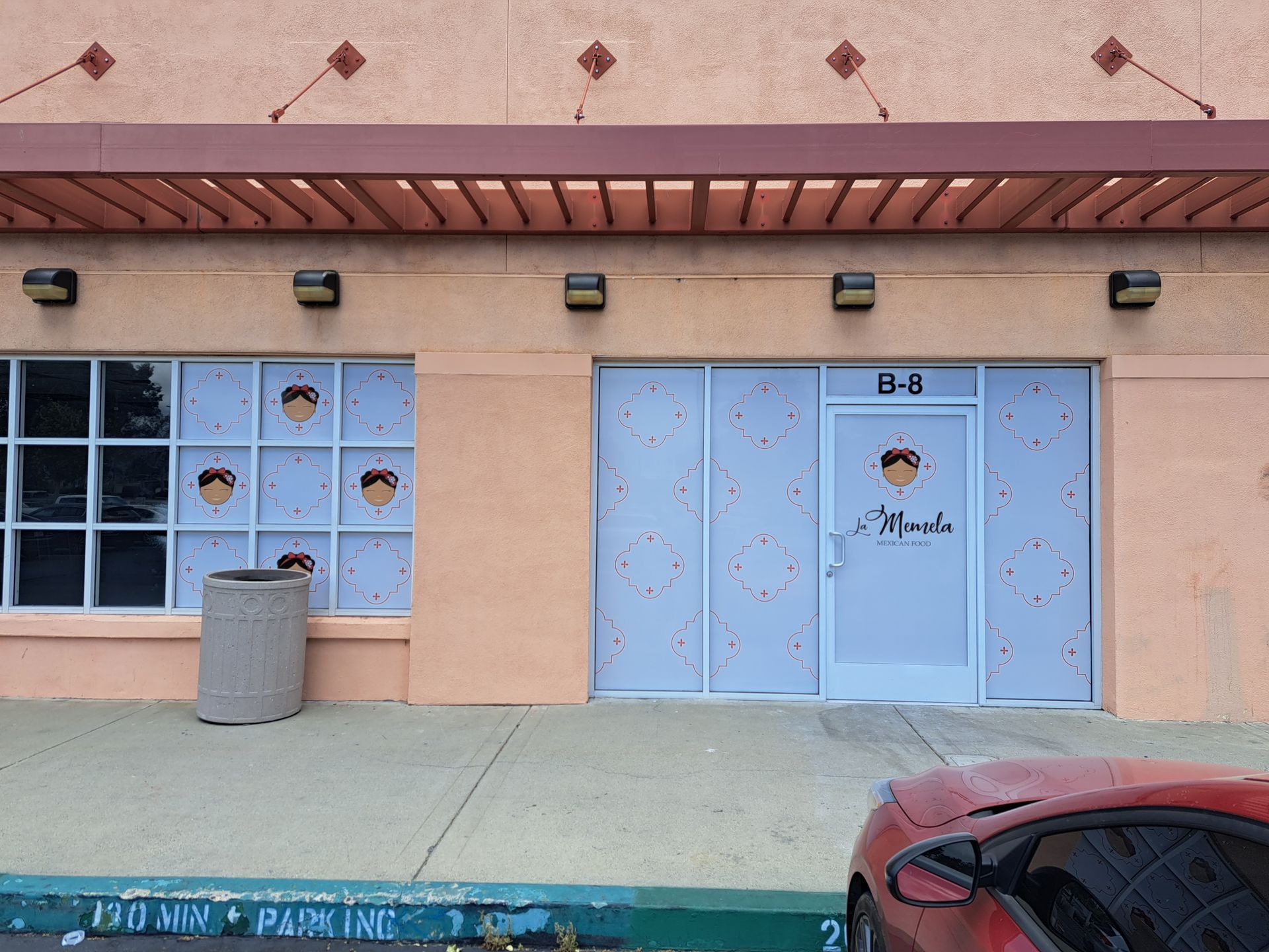 Storefront with blue double doors and windows, beige facade, trash can, and a red car in front.