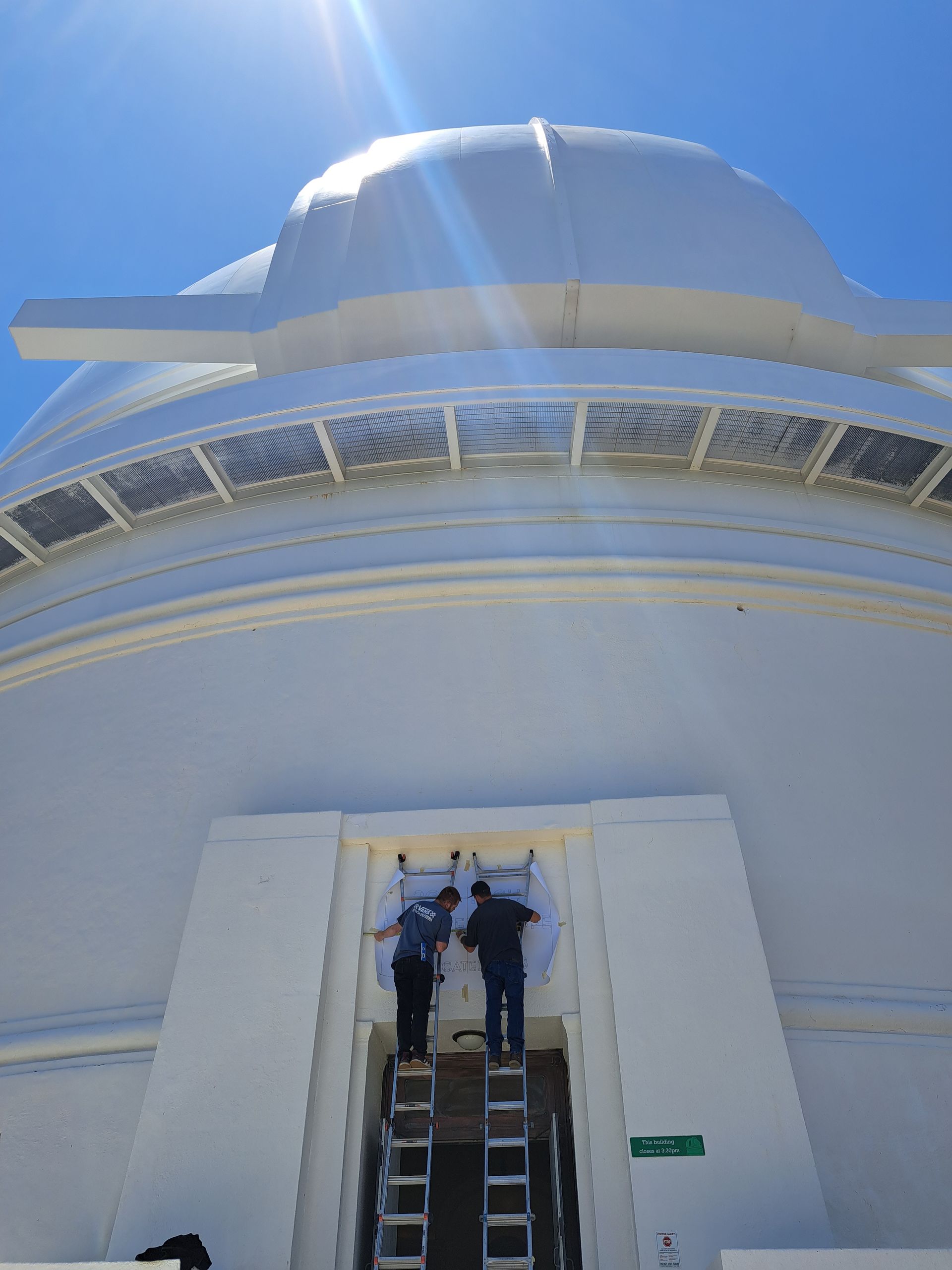 Two workers on a ladder painting or repairing a large white curved structure under a blue sky