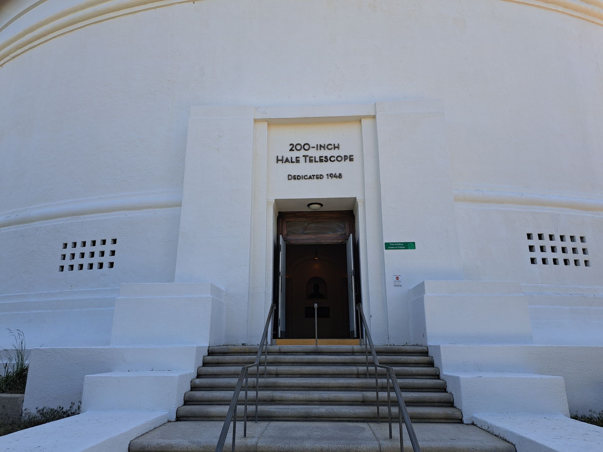 White museum entrance with steps and open doorway, framed by a curved facade and small windows