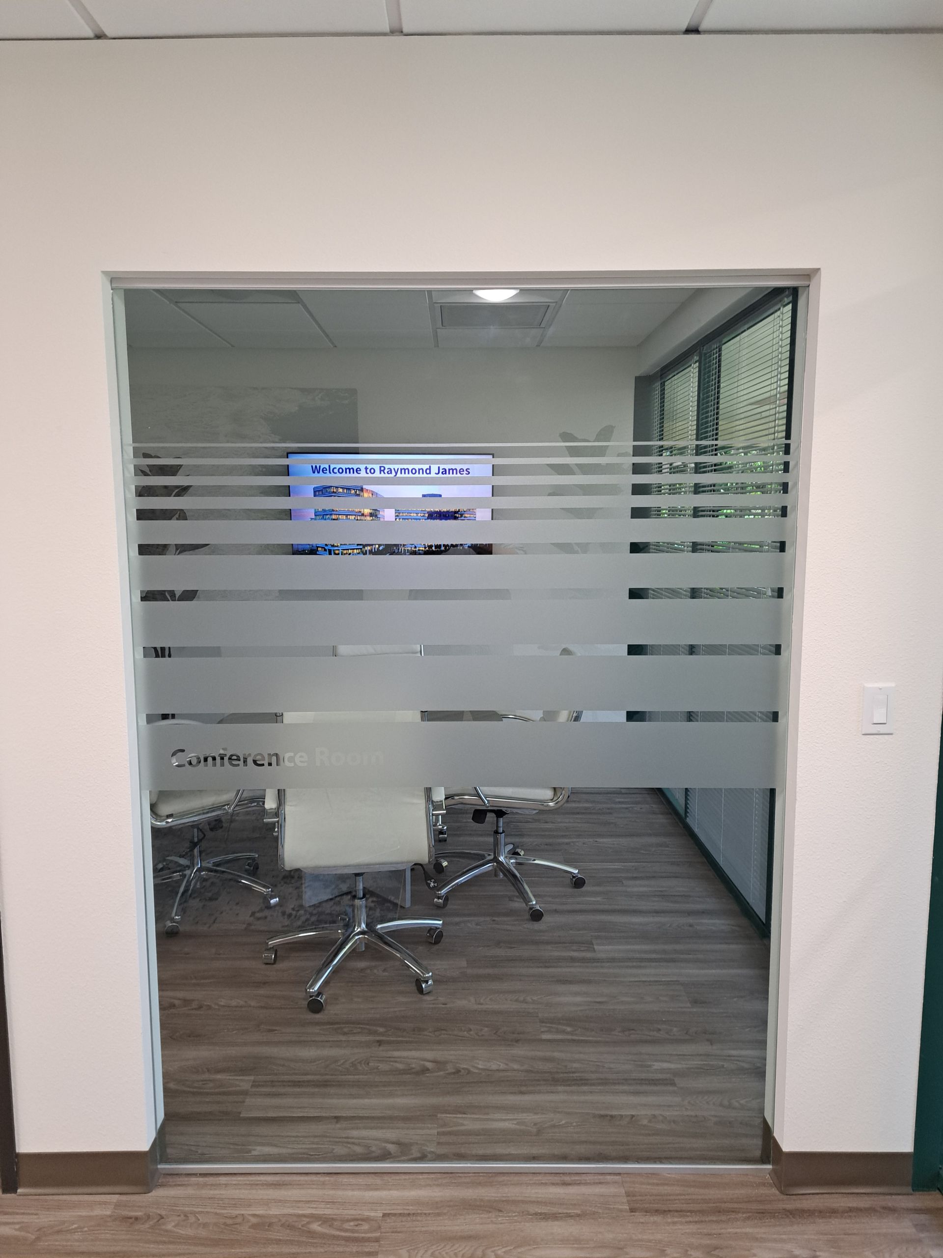 Glass-walled conference room with chairs and a wall-mounted screen inside a hallway.