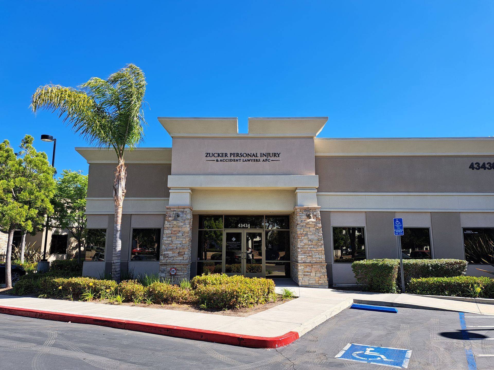 Strip mall storefront with stone columns, glass doors, and parking lot under a clear blue sky