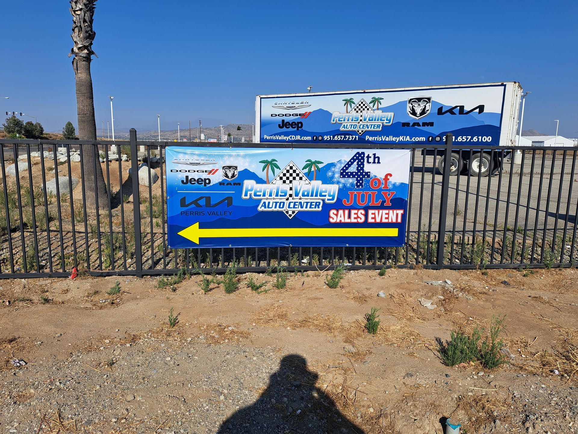 Blue event banners on a black fence by a beach, with a large yellow left arrow and a palm tree nearby.