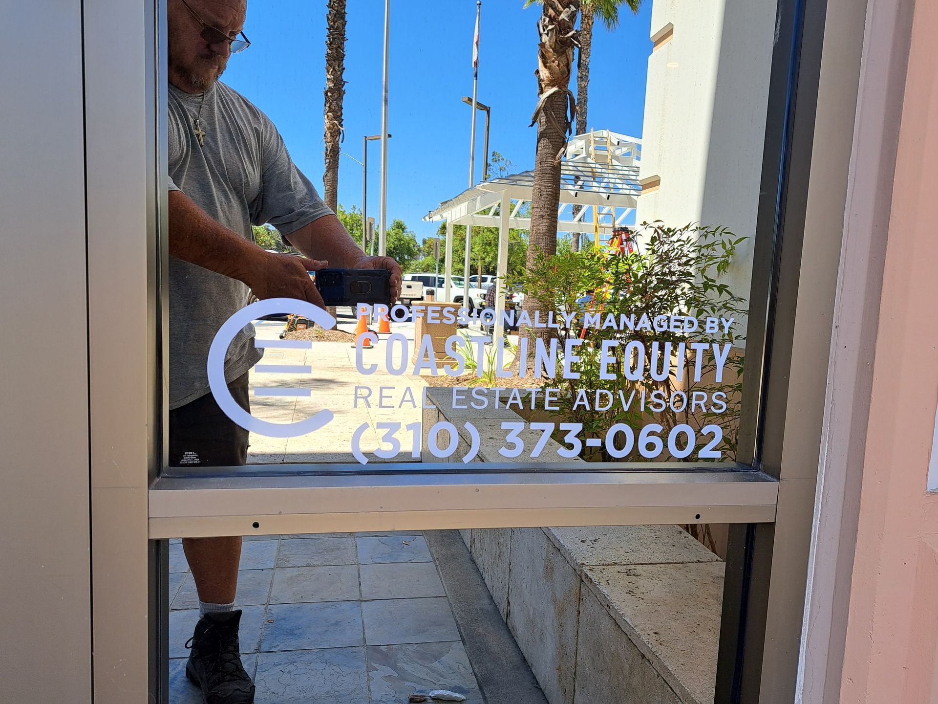 Glass door with white business lettering and phone number, reflecting a person opening it outside.