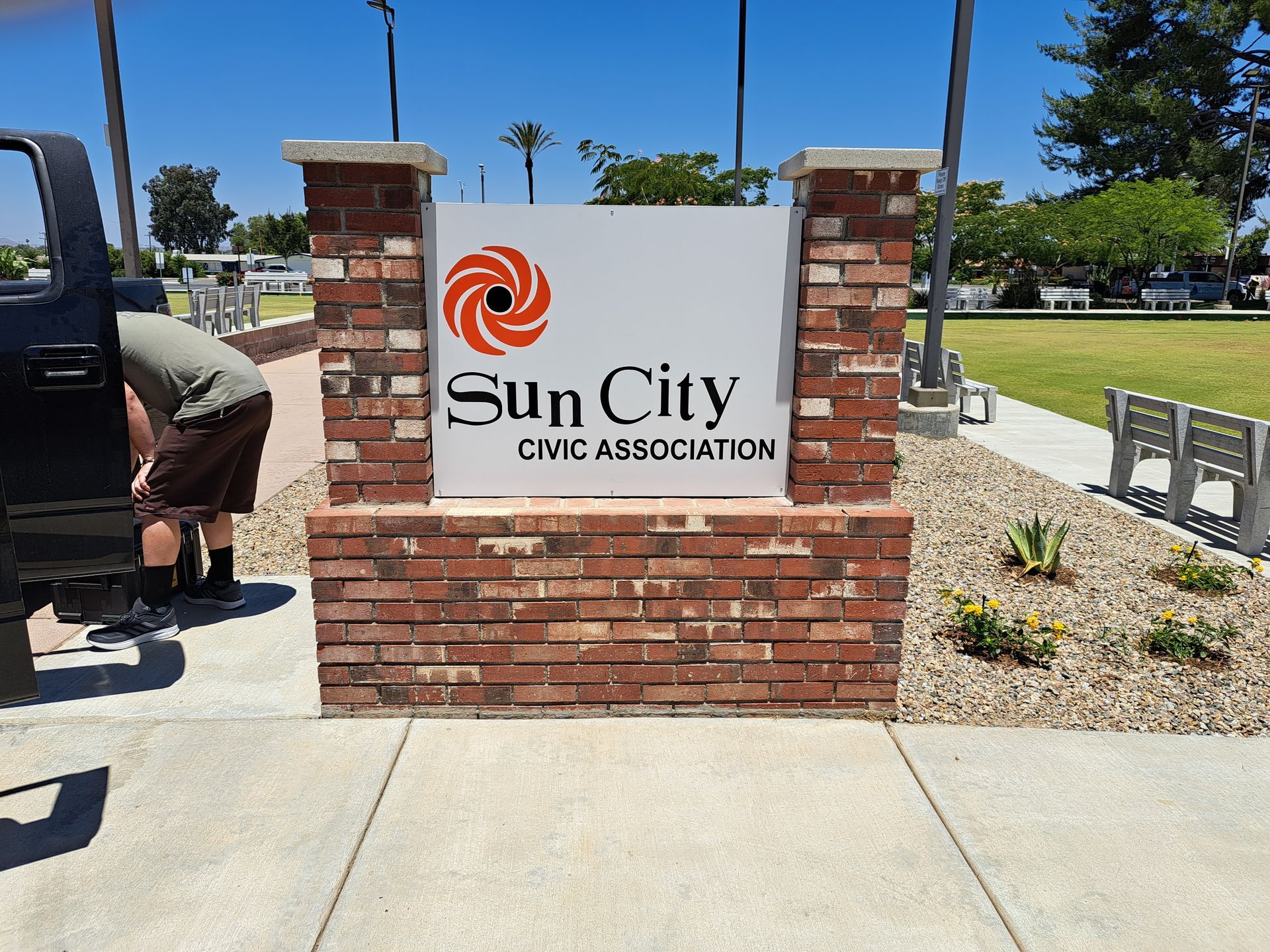 Sun City Civic Association sign on a brick wall beside a sidewalk and grassy park area