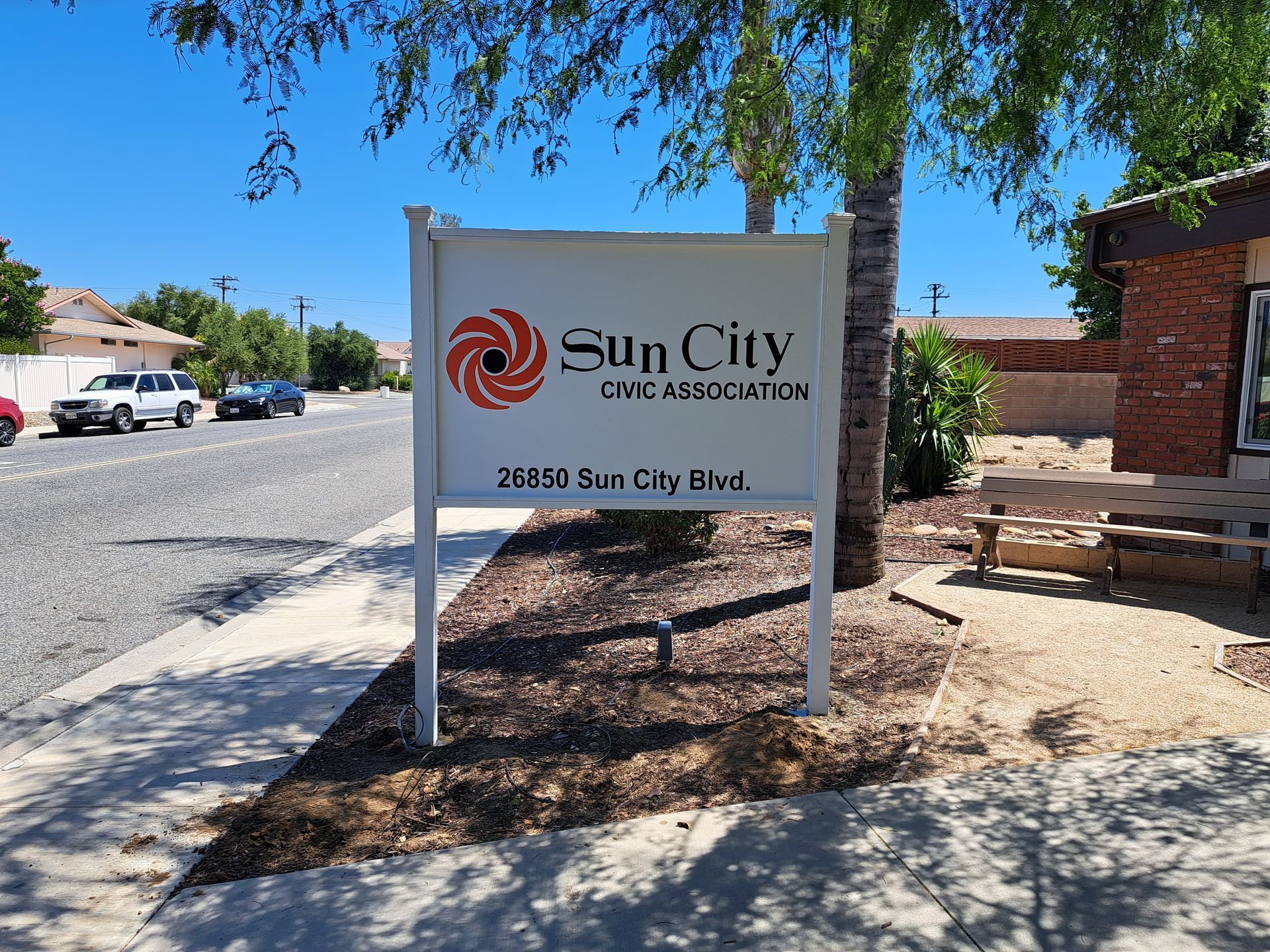 Sun City HOA entrance sign on a sunny street, with trees, parked cars, and a brick building nearby