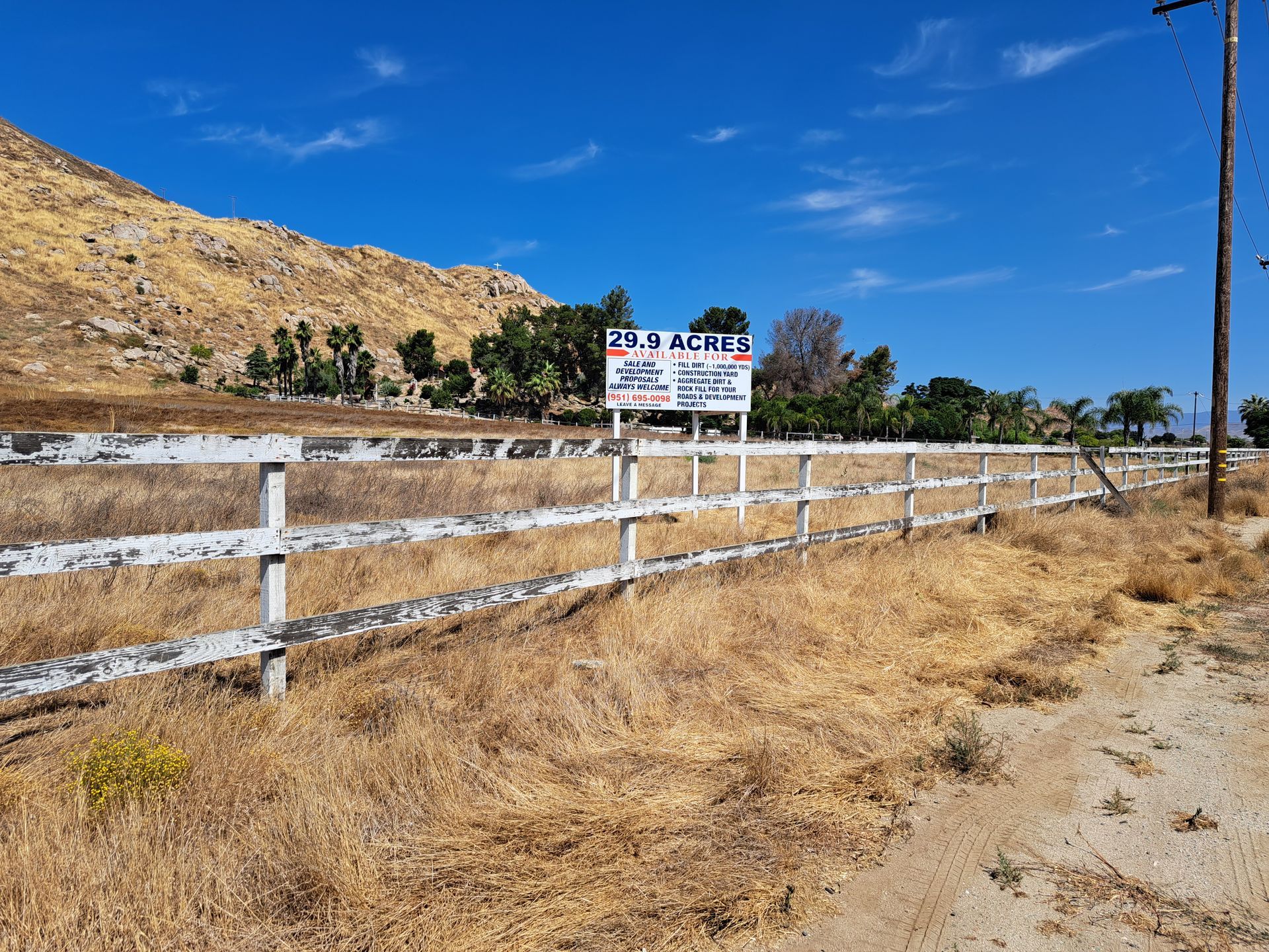 Dry rural roadside with white fence, dry grass, hills, and a small roadside sign under a blue sky