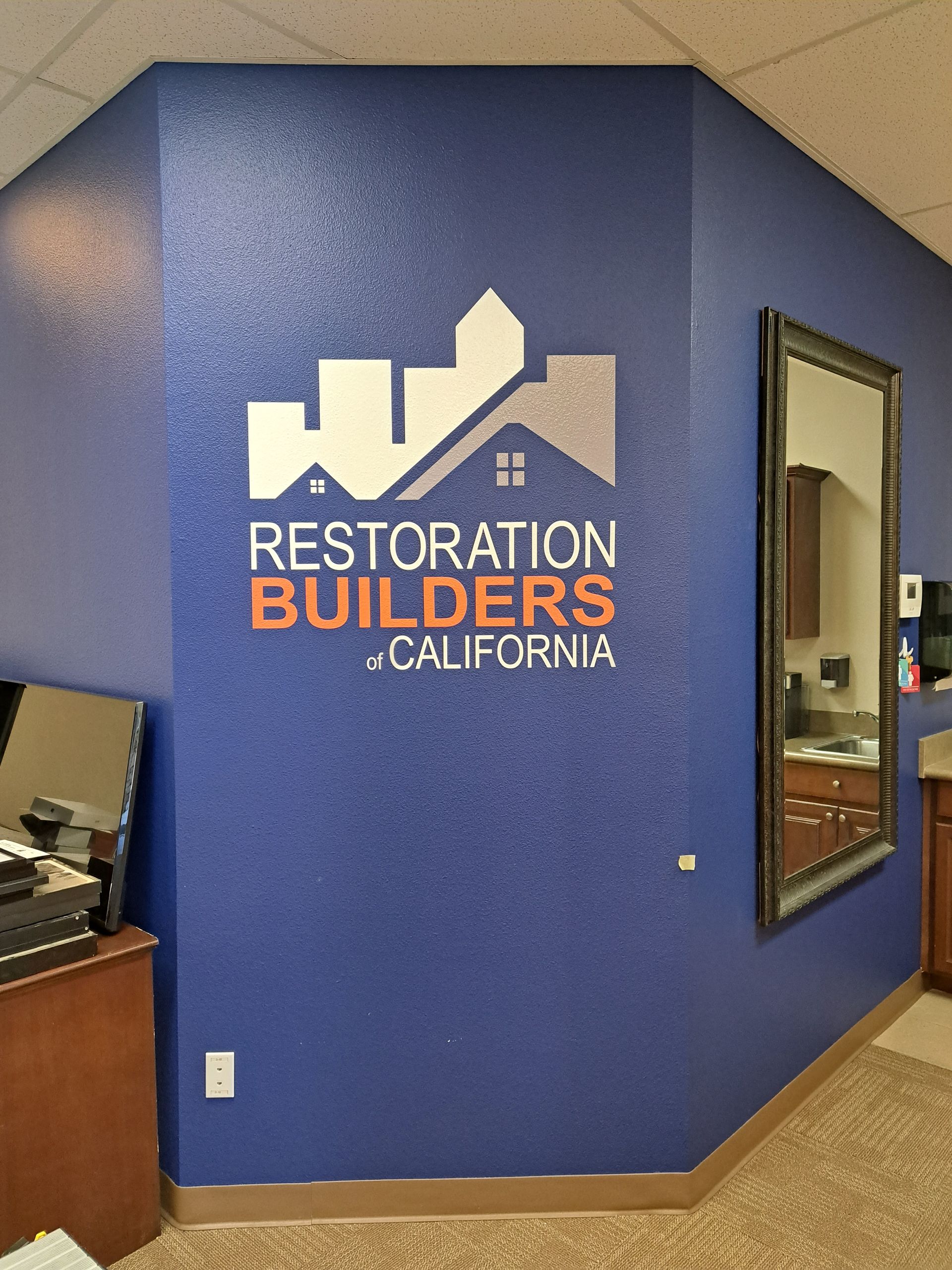Blue office wall with Restoration Builders California logo and a mirror on the right