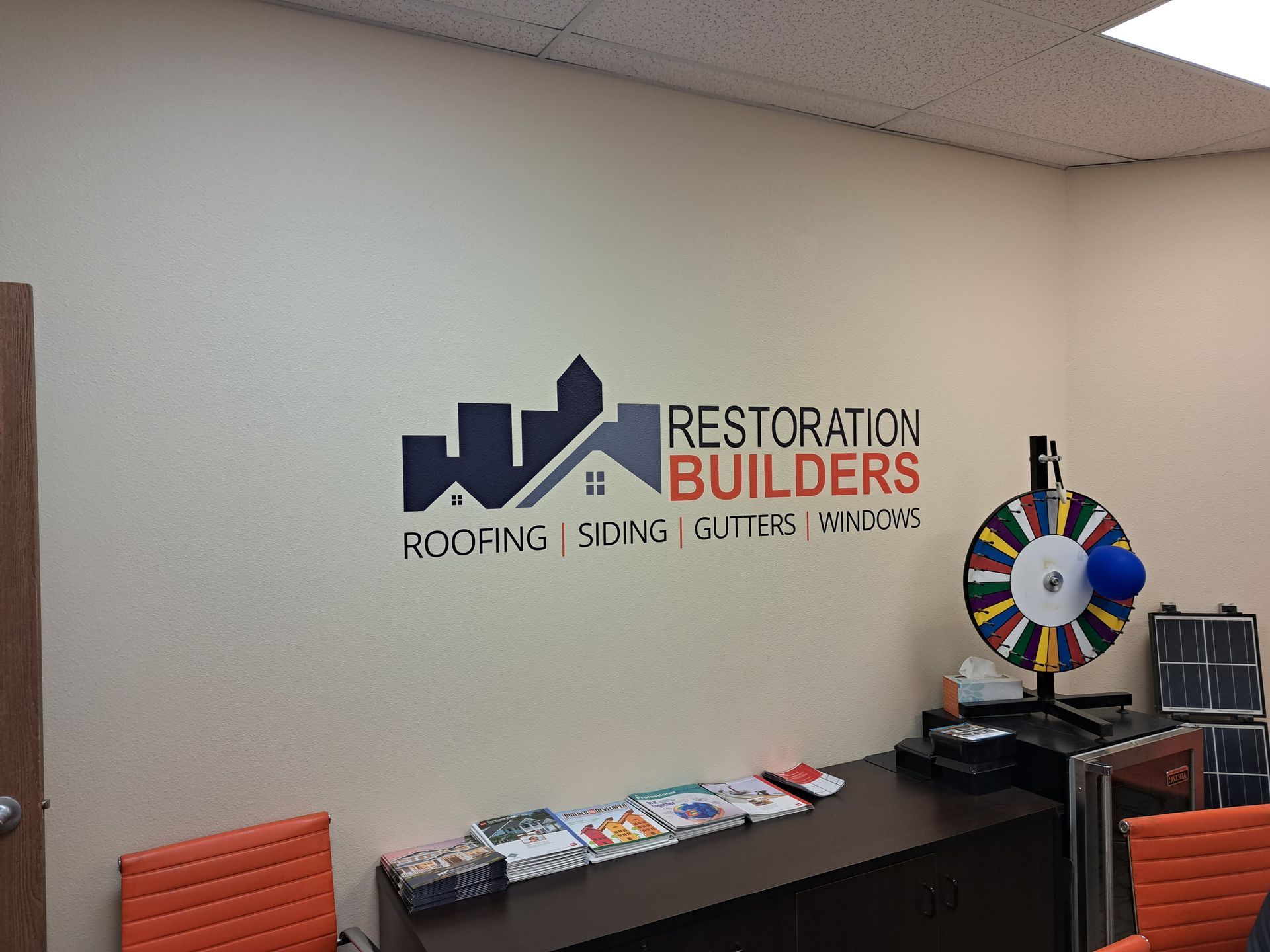 Office wall with Restoration Builders logo, black console table, and colorful prize wheel