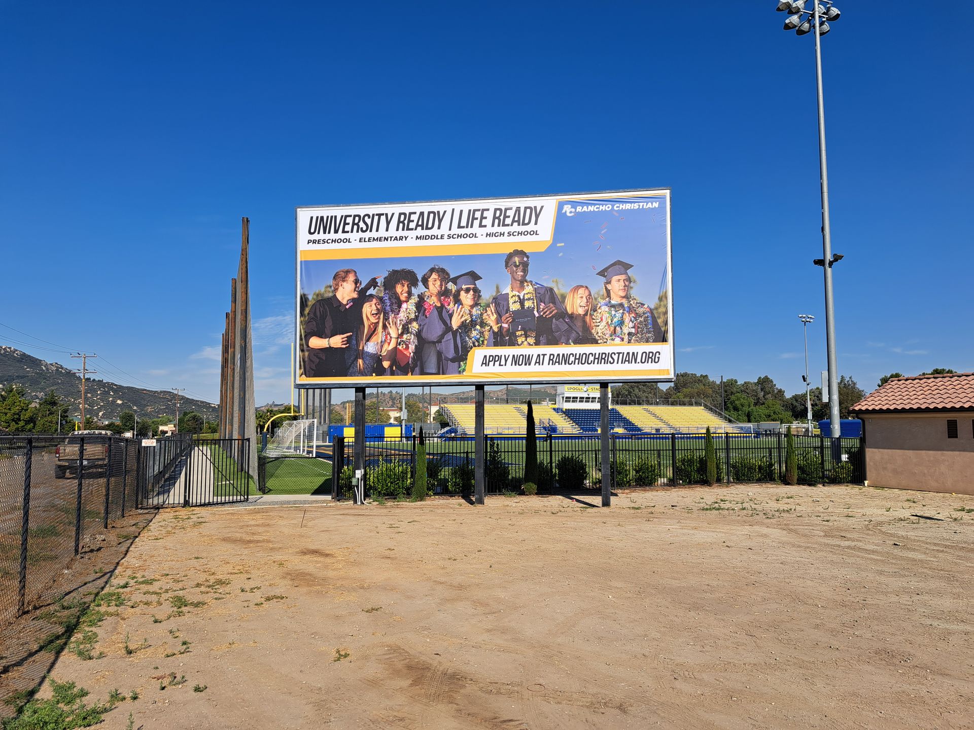 Outdoor billboard with a group photo and large text, set beside a dirt lot and fence under a clear blue sky