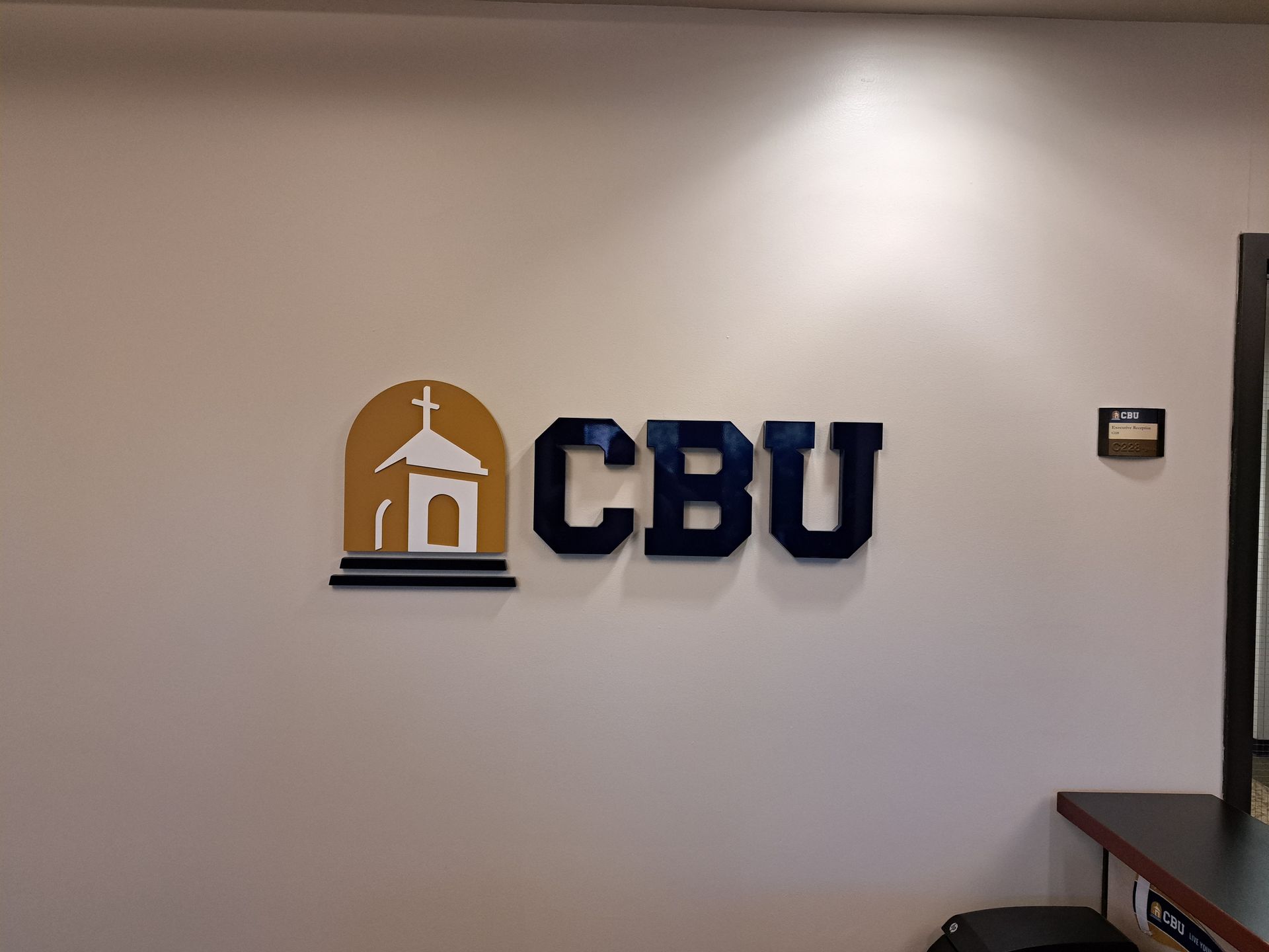 CBU logo on a white office wall, with a small cross icon in gold and blue lettering