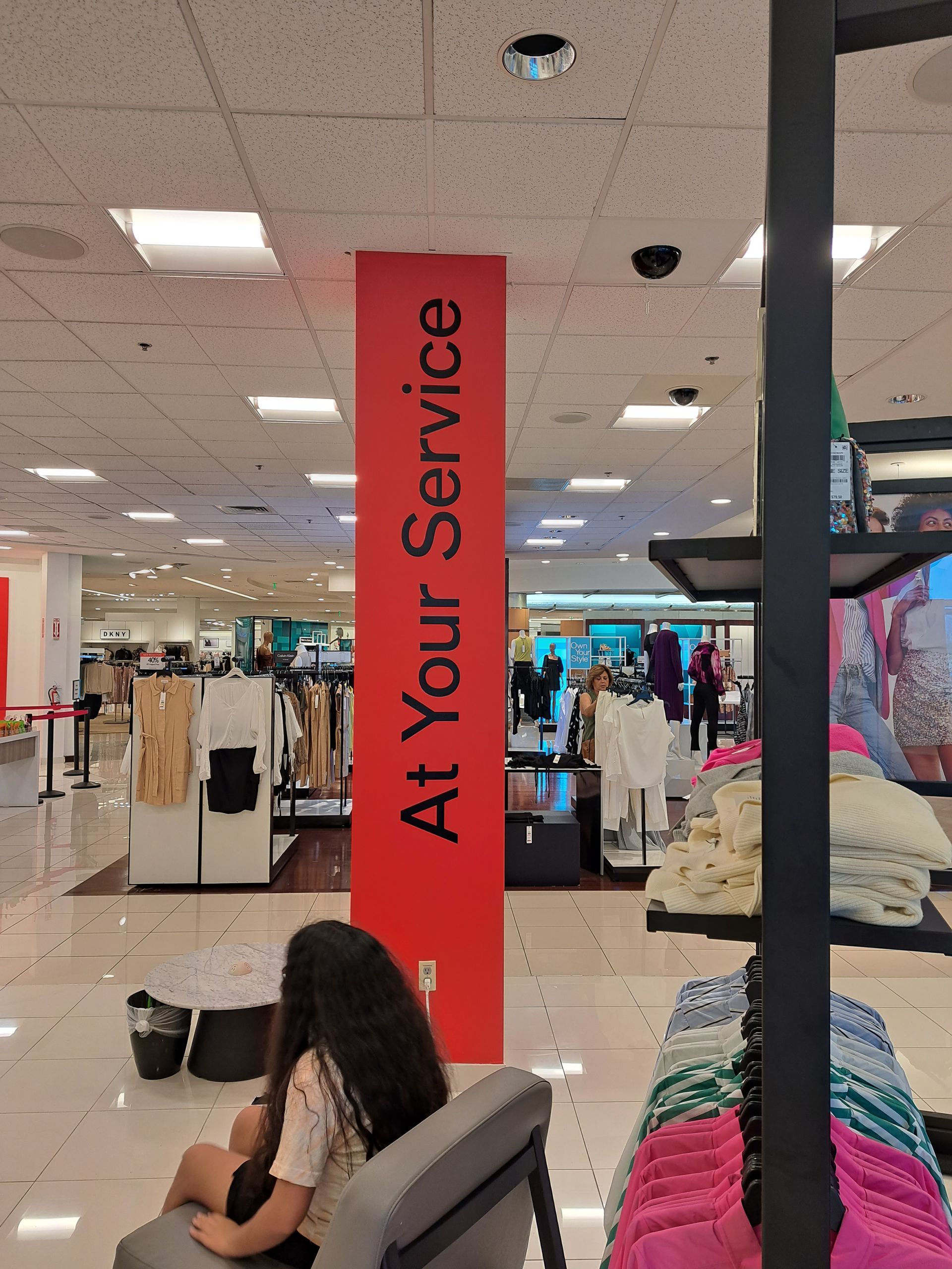 Retail store checkout area with a red “At Your Service” banner and shoppers seated nearby