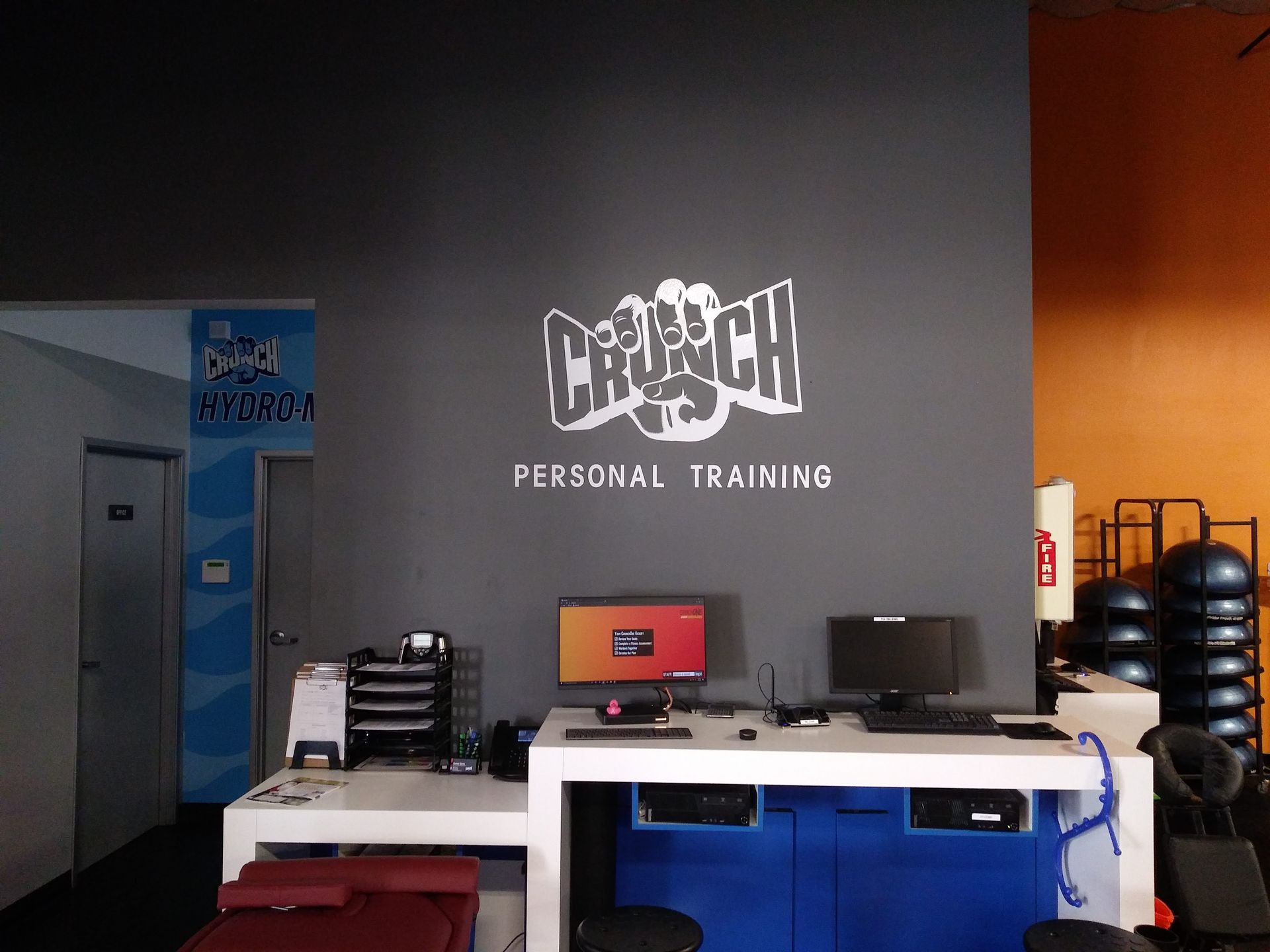 Gym reception area with a white desk, red laptop, and “CrossFit” logo on a dark wall.